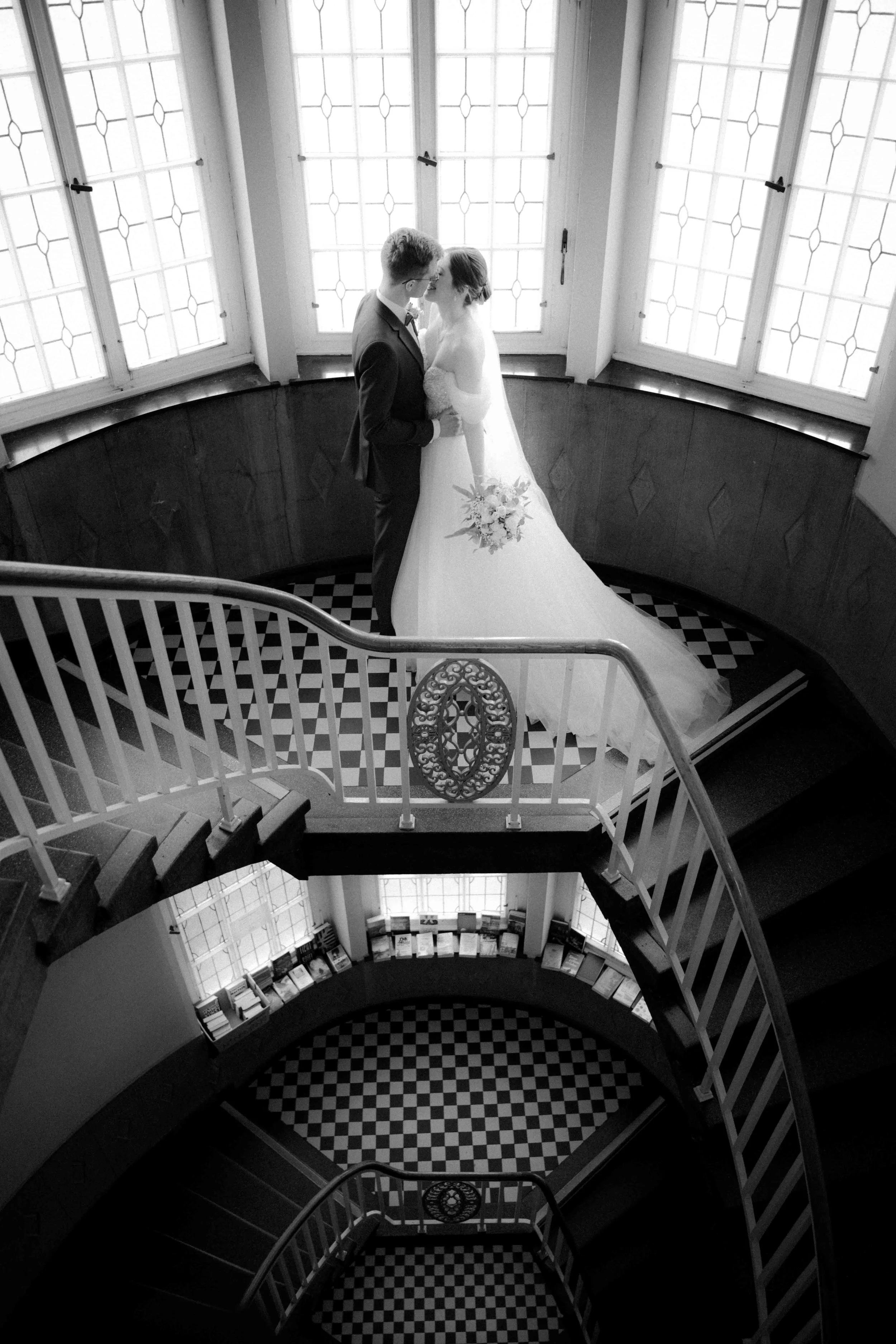 Ein schwarz-weiß-Foto eines Brautpaares, das sich auf einer Treppe in einem alten Gebäude küsst, vor großen Fenster mit Sprossen. Die Braut trägt ein langes weißes Kleid und hält einen Blumenstrauß, der Bräutigam trägt einen dunklen Anzug. Die Hochze