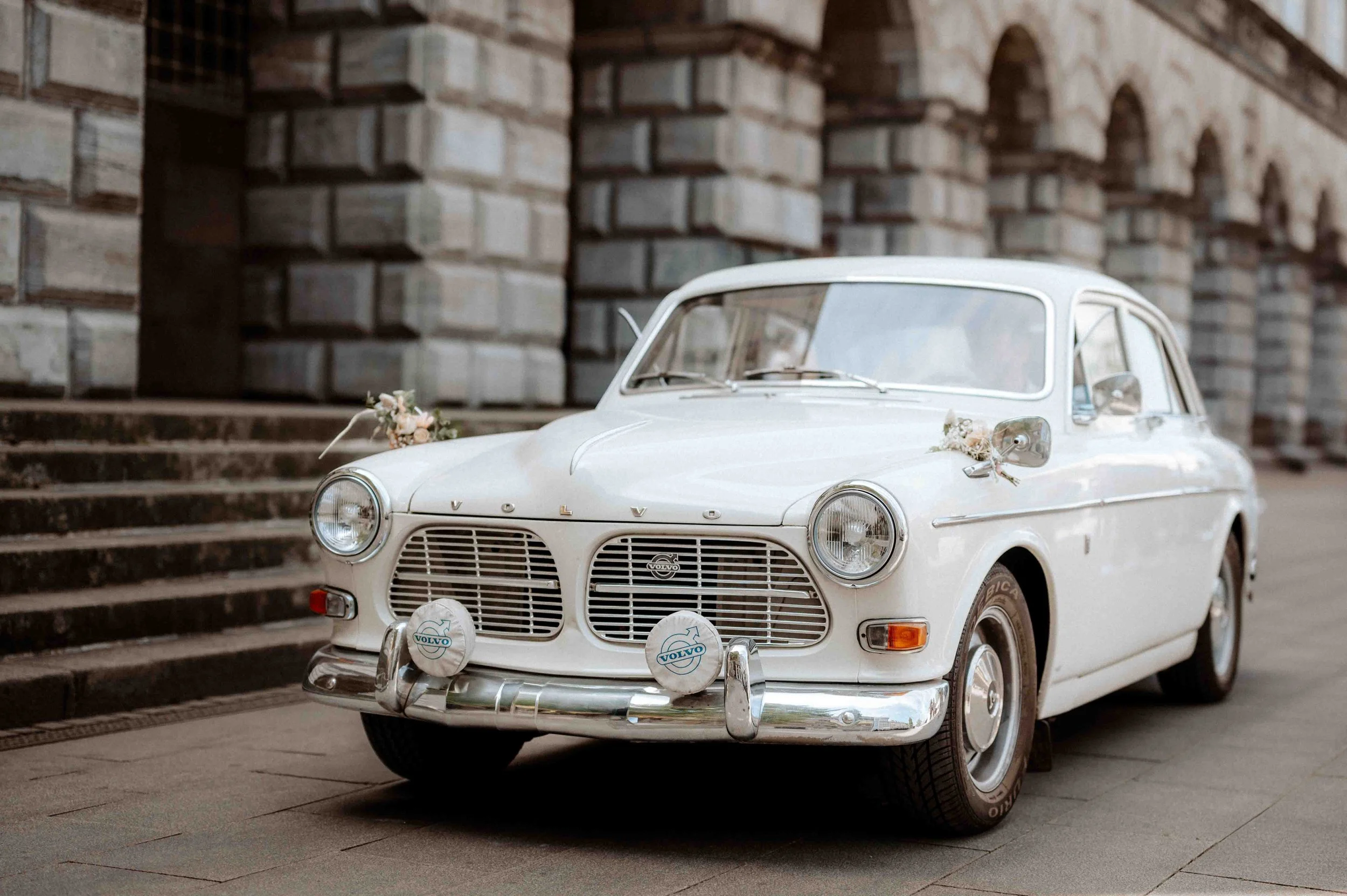 Ein weißer, vintage Volvo Autos auf einer Straße, verziert für eine Hochzeit, mit Blumen und Schutzabdeckungen auf den Scheinwerfern. Die Hochzeit findet im Standesamt in Mülheim an der Ruhr bei Düsseldorf statt. 