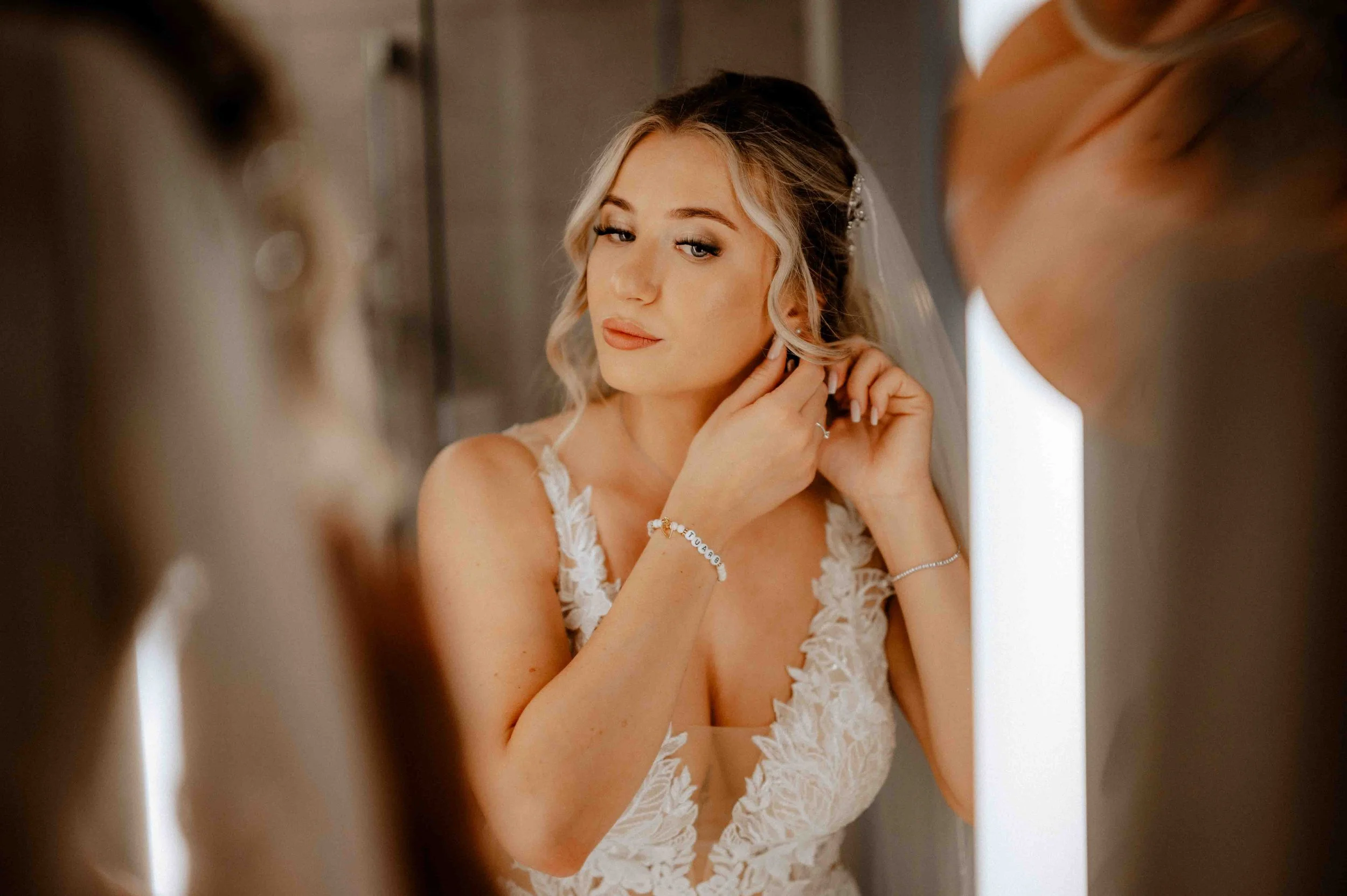 Junge Frau in Brautkleid schaut in den Spiegel, während sie sich einkleidet und ihre Ohrringe anlegt. Die Hochzeit findet in Köln statt. Es handelt sich um das Getting Ready der Braut vor der Hochzeit. 