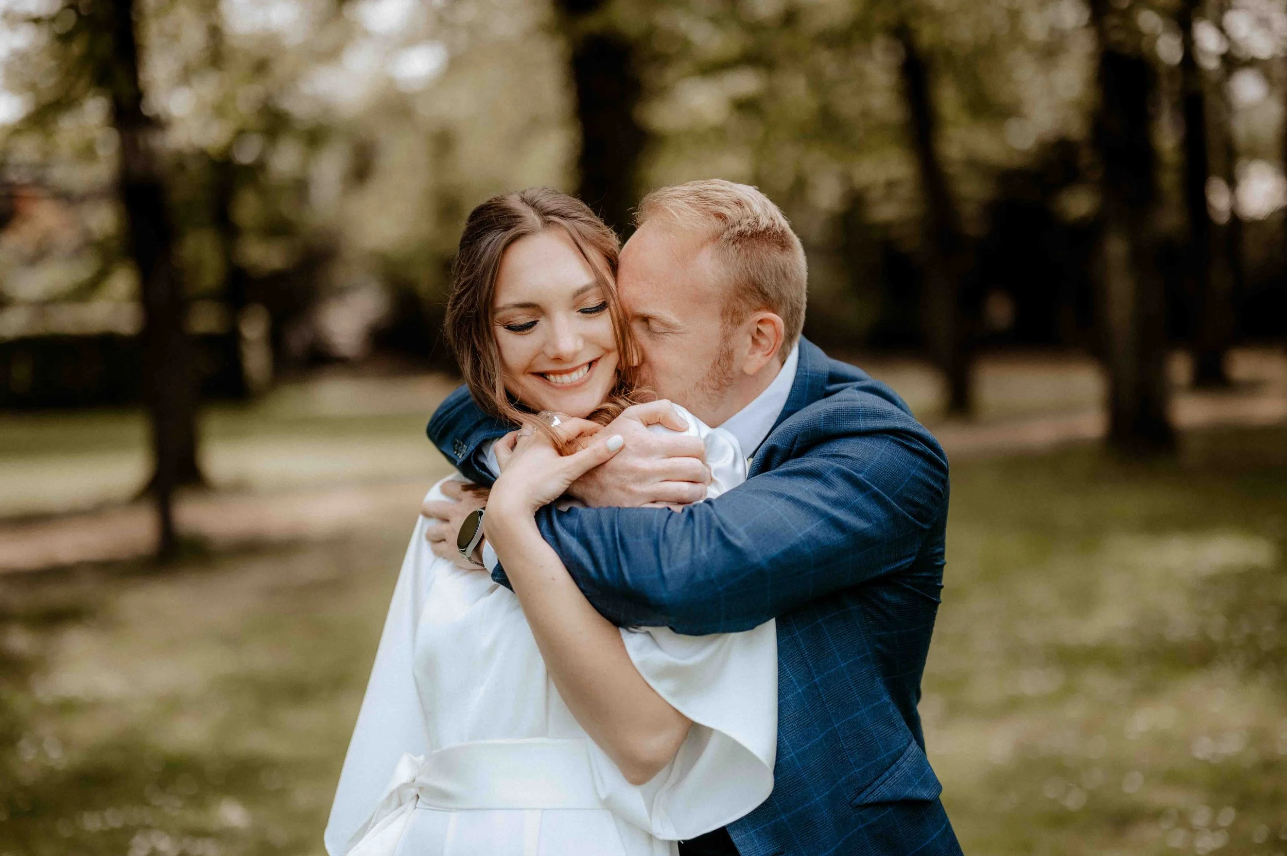 Ein glückliches Paar umarmt sich in einem Park, im Hintergrund Bäume, bei Tageslicht. Die Hochzeit findet im Haus Cromford in Ratingen bei Düsseldorf, Nähe Köln statt. 