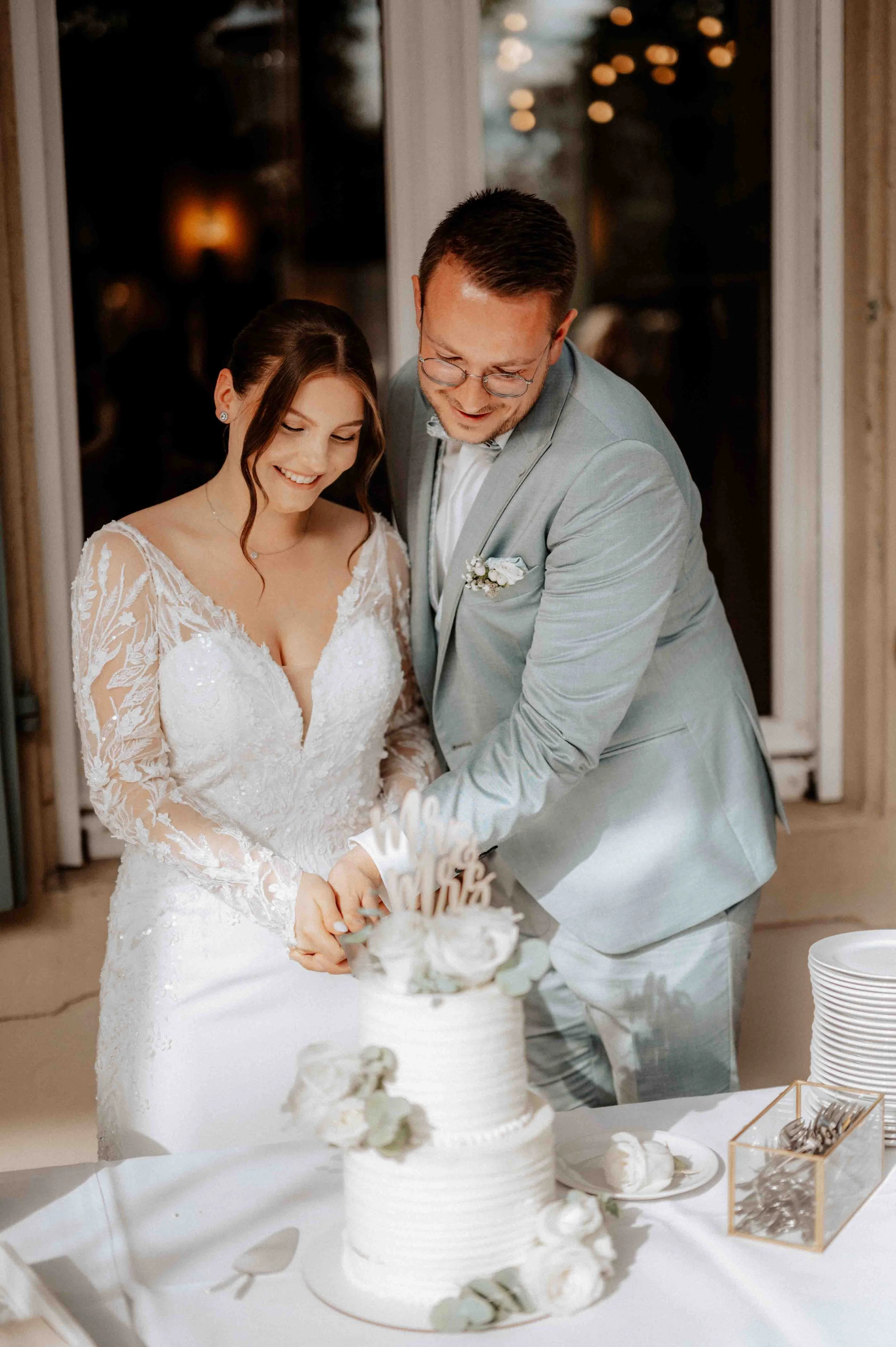 Ein Brautpaar schneidet gemeinsam eine Hochzeits-Torte bei ihrer Feier, im Hintergrund festliche Beleuchtung. Die Hochzeit findet im Schloss Eicherhof in Leichlingen, Nähe Köln und Düsseldorf statt. 