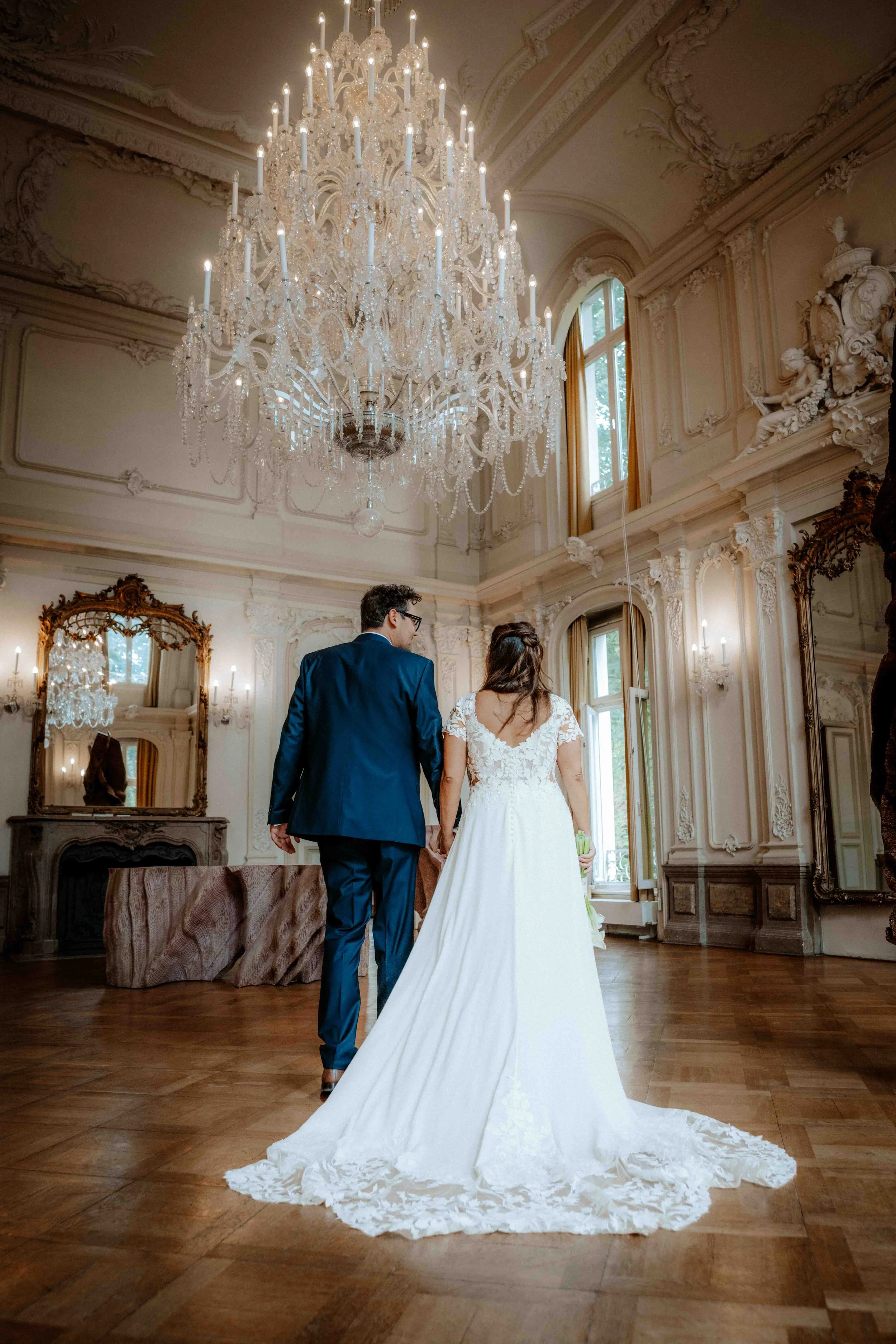 Braut und Bräutigam halten Händchen in einem eleganten, historischen Raum mit Kronleuchter, großen Fenstern und ornamentalen Wandverzierungen. Die Hochzeit findet im Museum bzw. Schloss Morsbroich in Leverkusen, Nähe Köln statt. 