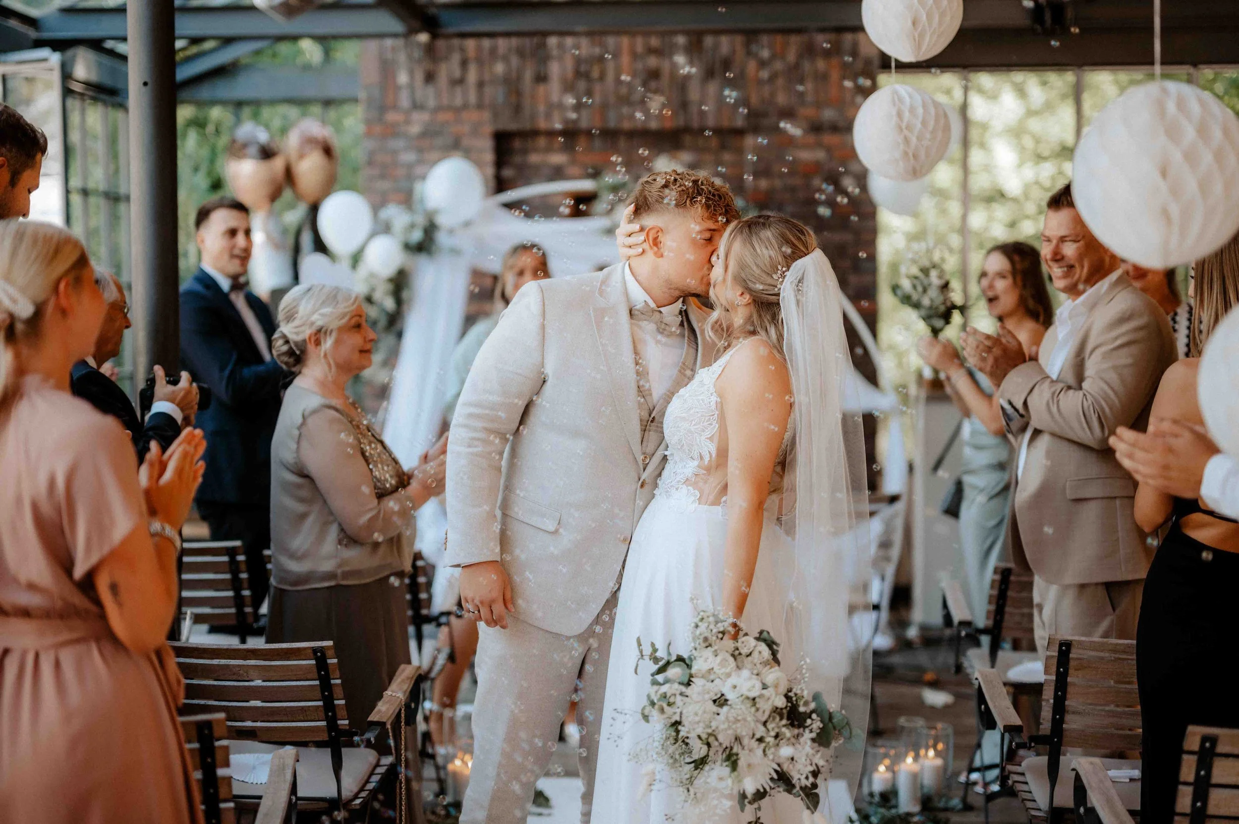 Ein frisch verheiratetes Paar teilt sich einen Kuss während ihrer Hochzeitszeremonie, umgeben von applaudierenden Gästen in einem festlich dekorierten Innenraum. Die Hochzeit findet in Bergisch Gladbach bei Köln statt. 