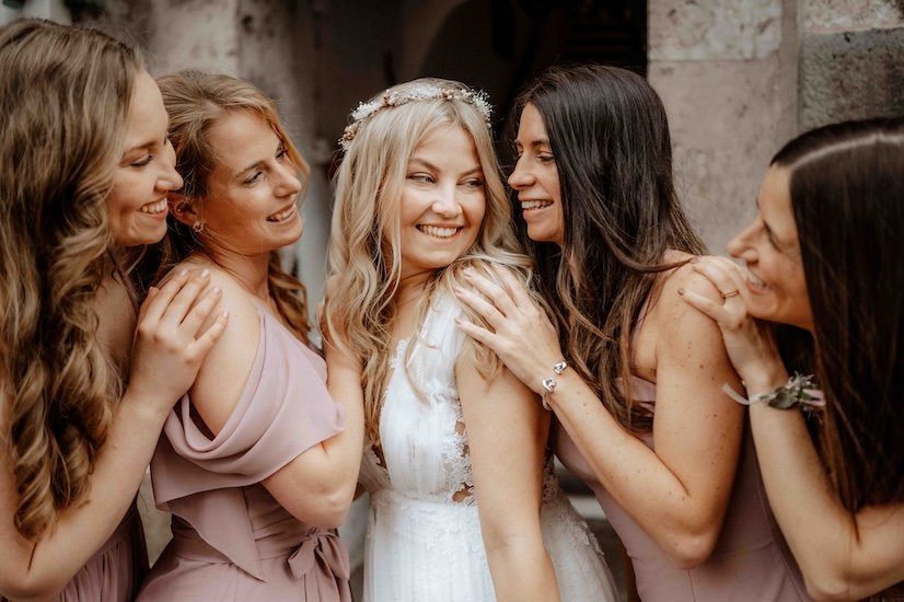 Hochzeitsszene mit fünf lachenden Frauen, eine Braut in weißem Kleid und blondem Haar, umgeben von vier Bräuten in lilafarbenen Kleidern, die sich zusammen freuen und lachen. Die Hochzeit findet in der Severinstorburg in Köln statt.