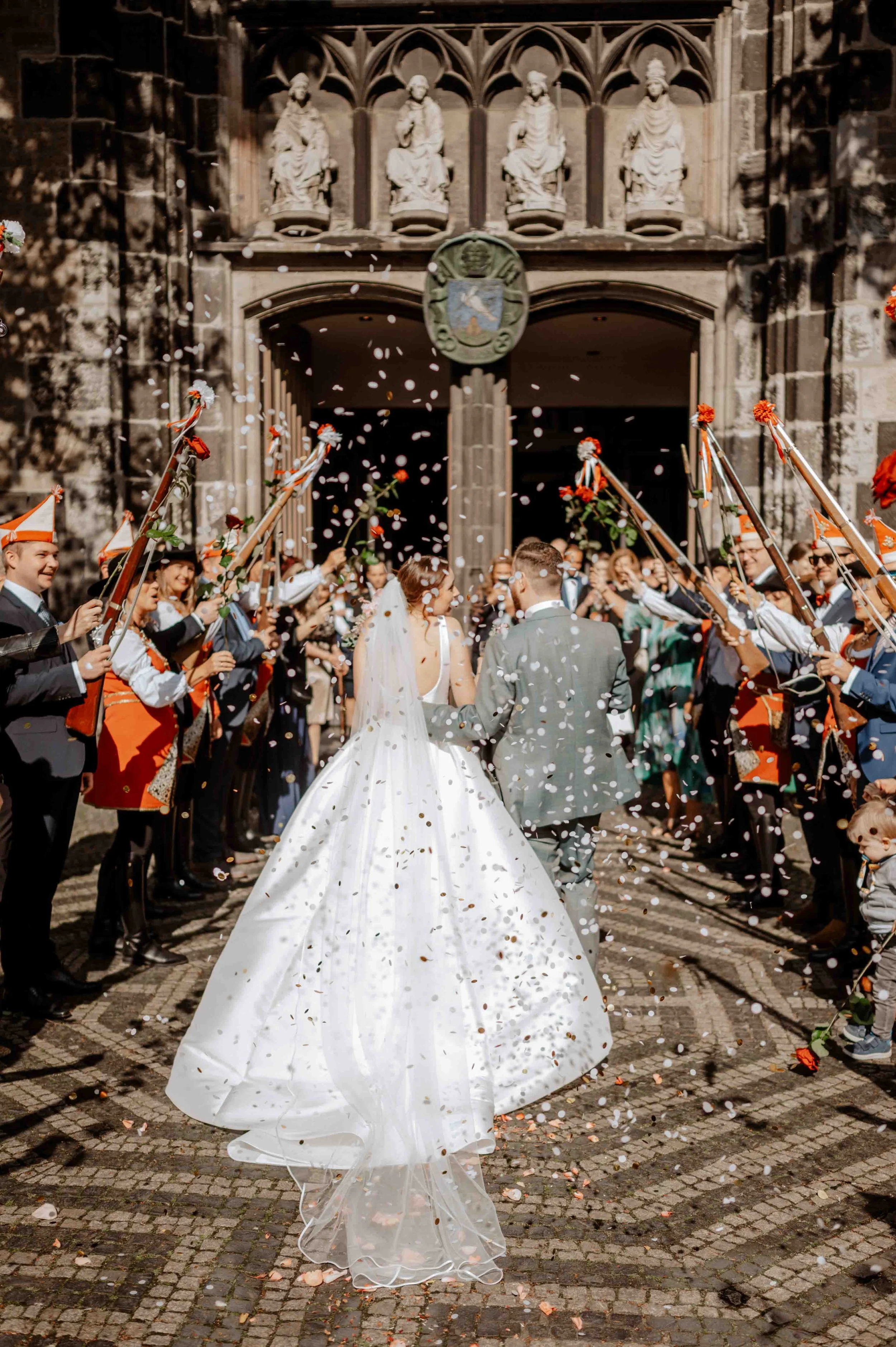 Ein frisch verheiratetes Paar wird vor einer historischen Kirche gefeiert, während Gäste Konfetti werfen. Die Hochzeit findet in Köln in der Kirche Sankt Severin statt. 