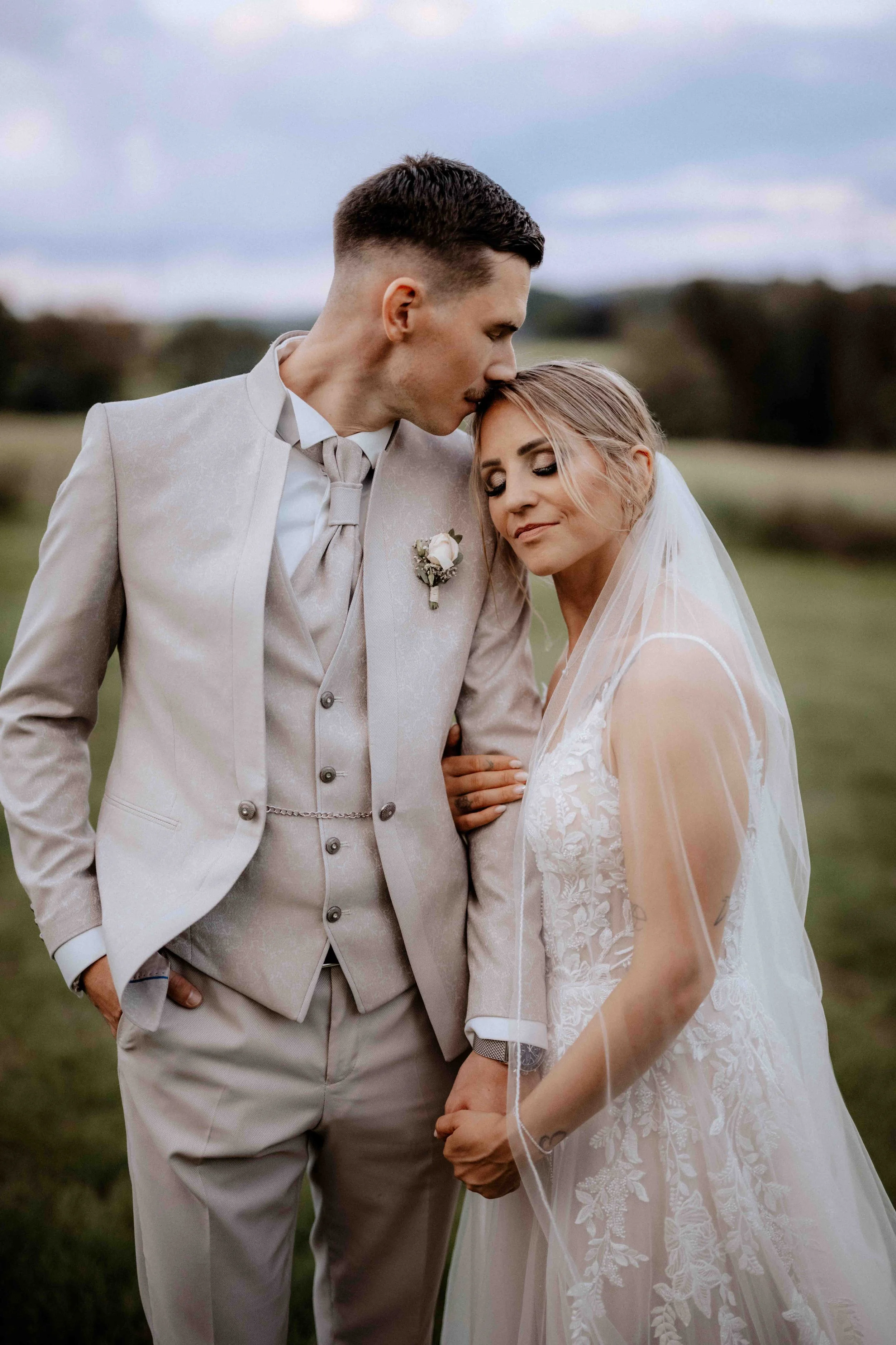 Braut und Bräutigam arm in arm in einer weiten Wiese, bei Sonnenuntergang, elegante Hochzeitskleidung, zärtlich küssend, emotionaler Moment. Die Hochzeit findet in Düsseldorf, Nähe Köln statt. 