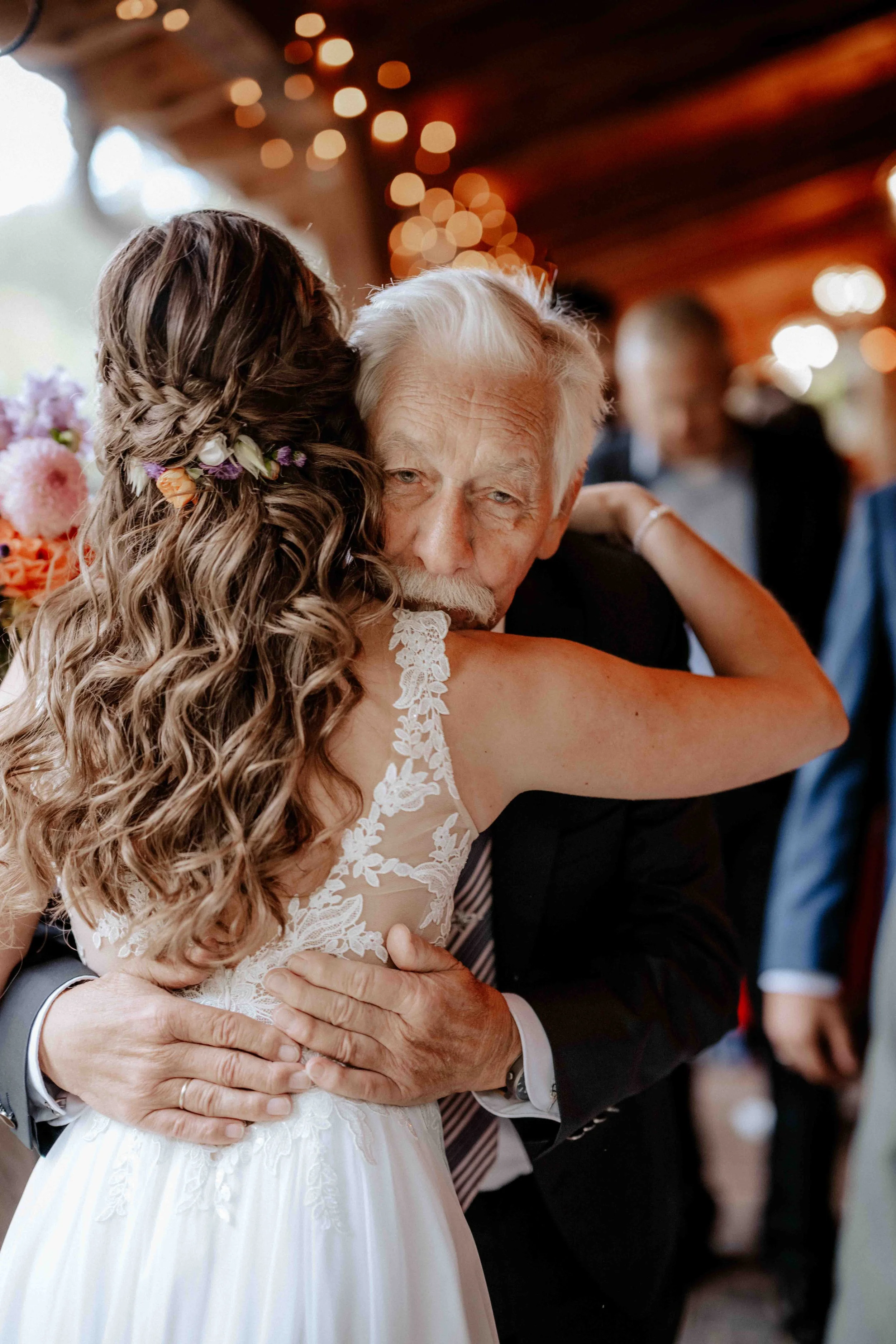 Ein älterer Mann umarmt eine junge Braut bei einer Hochzeitsfeier, umgeben von anderen Gästen und warmer Beleuchtung. Die Hochzeit findet in Nümbrecht bei der Holsteins Mühle, Nähe Köln statt. 