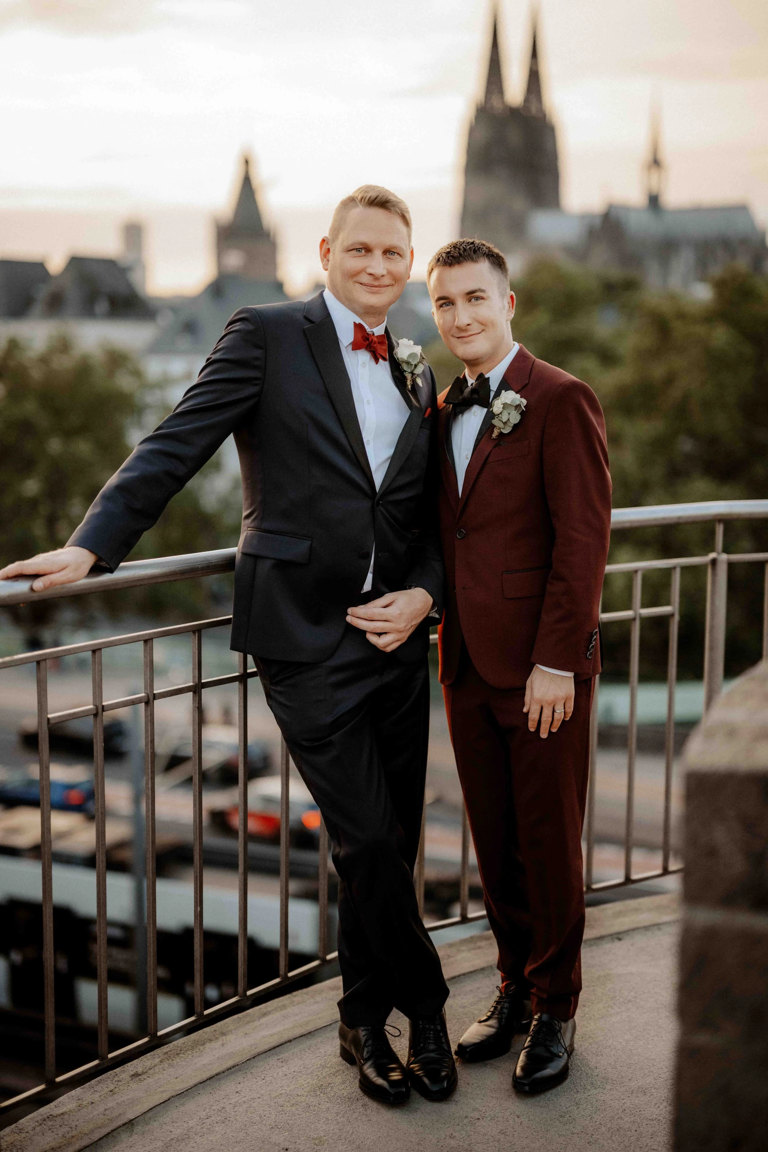 Zwei Männer in Anzügen auf einem Balkon, die sich anlächeln, mit einer Stadt im Hintergrund bei Sonnenuntergang. Die Hochzeit findet in Köln im Maritim Hotel statt. Im Hintergrund ist der Kölner Dom zu sehen. Es ist ein same sex Ehepaar.  