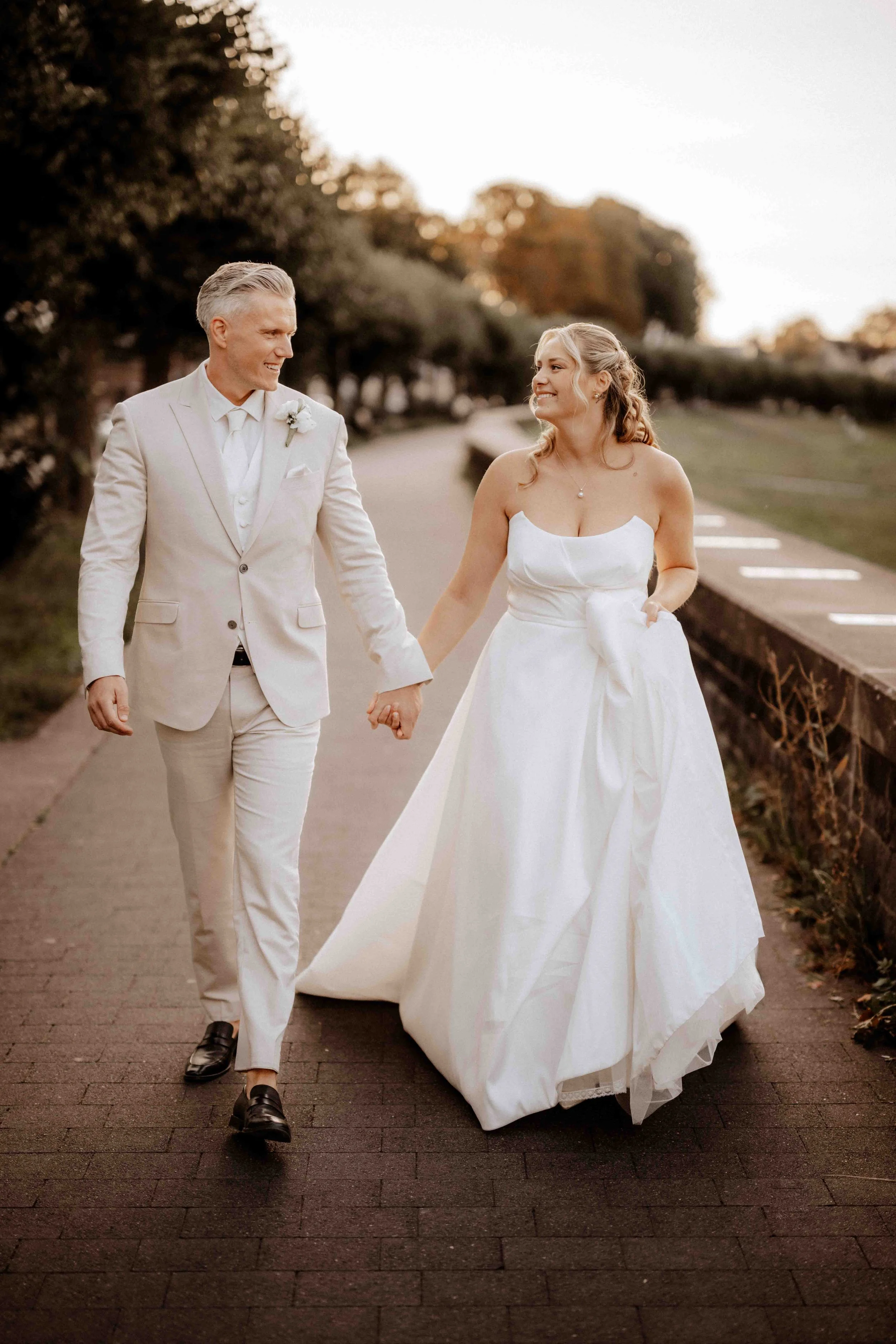 Ein Brautpaar in Hochzeitskleidung, beide lächelnd, gehen Hand in Hand auf einem gepflasterten Weg im Freien, umgeben von Bäumen bei Sonnenuntergang. Die Hochzeit findet in Köln Rodenkirchen am Rhein statt. 