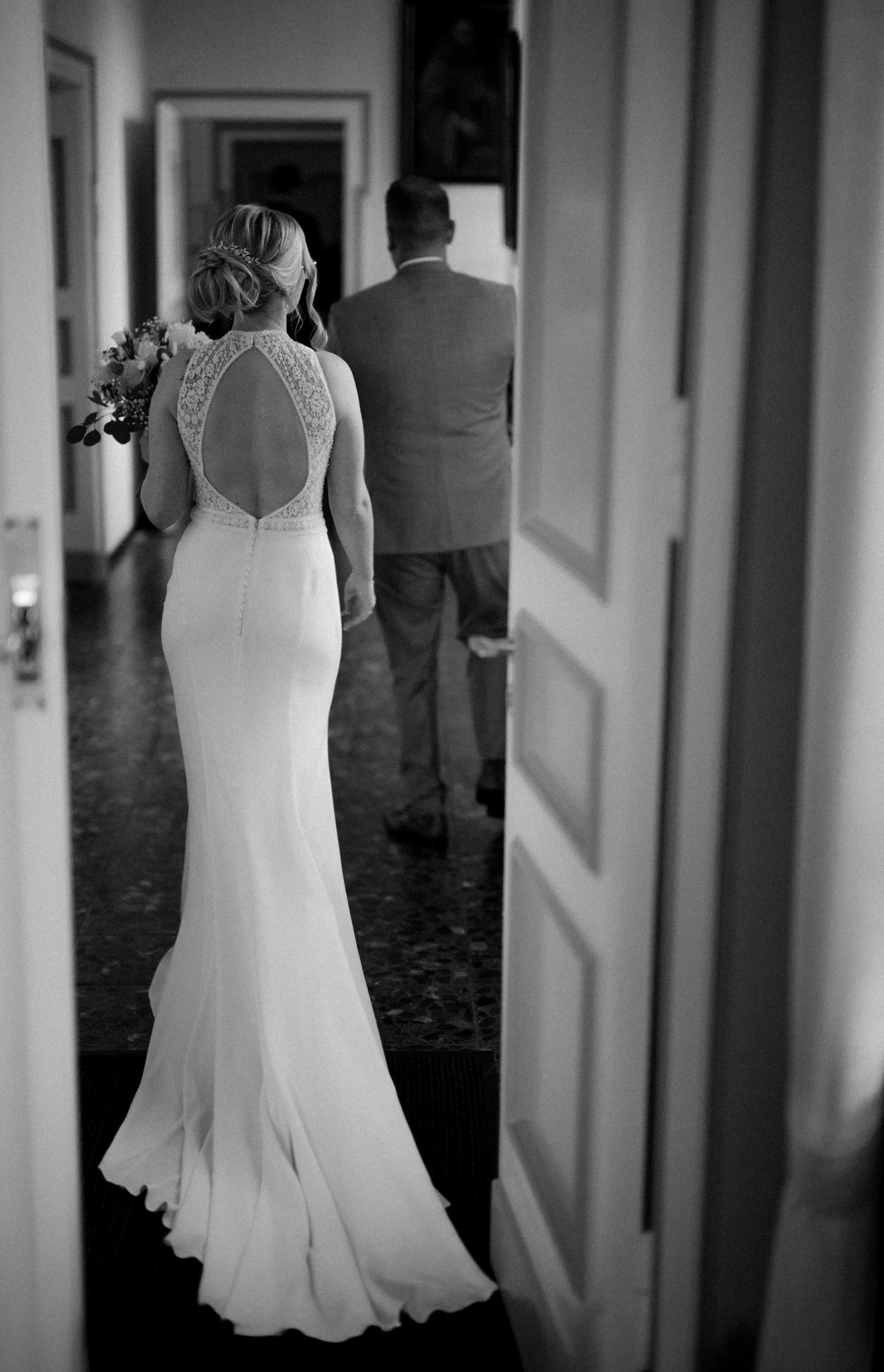 Braut in Hochzeitskleid mit Blumenstrauß, die einem Mann in Anzug folgt, beim Betreten eines Raums, schwarz-weiß Aufnahme. Die Hochzeit findet in Düsseldorf, Nähe Köln statt. 