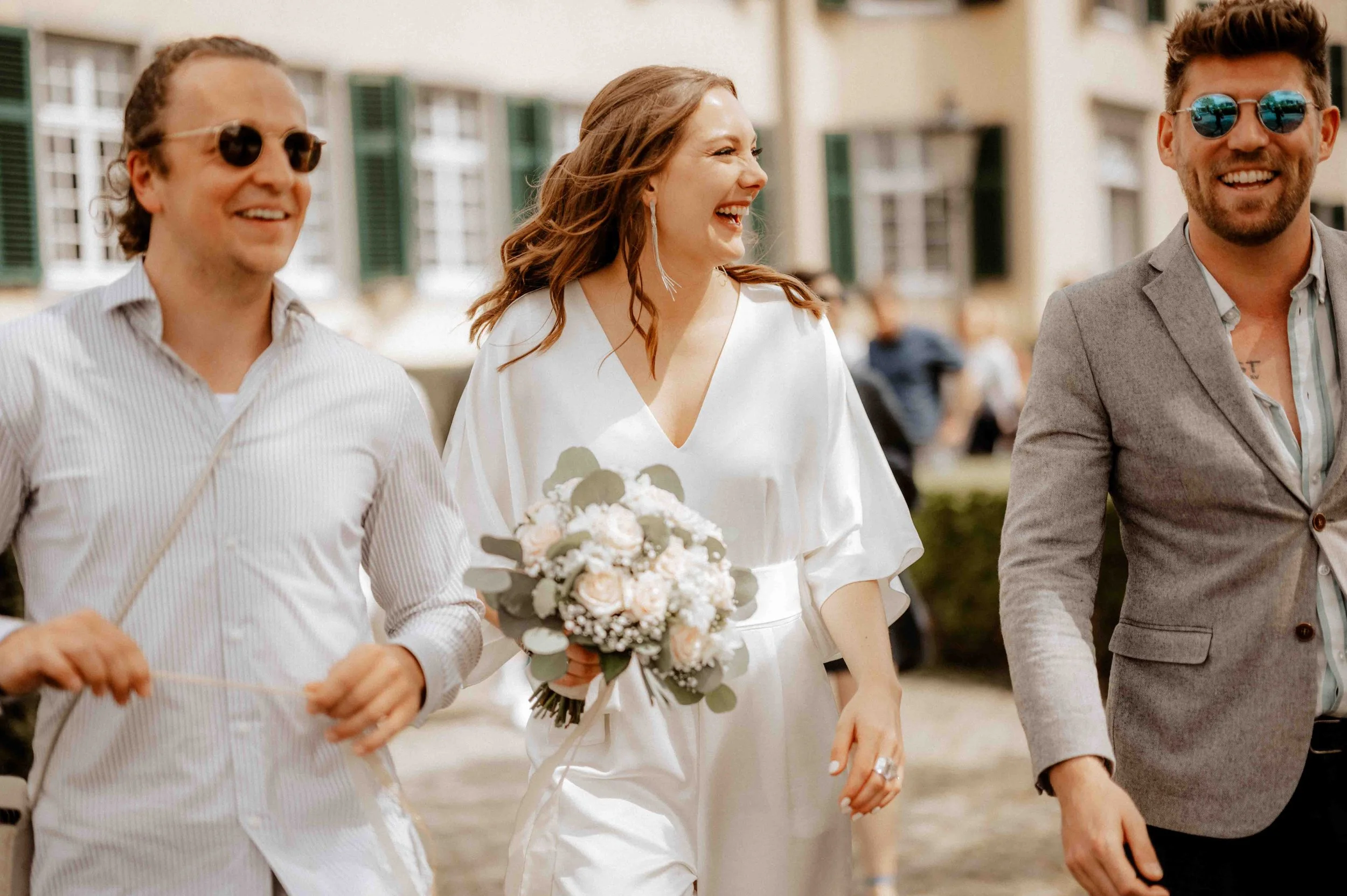 Drei Personen, eine Frau in weißem Kleid mit Blumenstrauß, lächelt und geht zwischen zwei Männern in Anzügen, die ebenfalls lachen, auf einer belebten Straße in einer Stadt. Die Hochzeit findet im Haus Cromford in Ratingen bei Düsseldorf statt. 