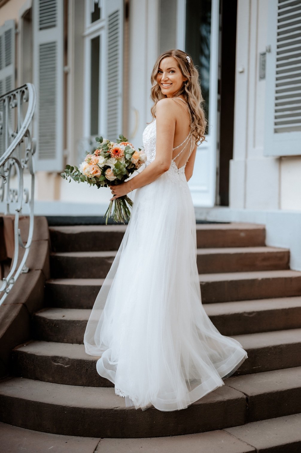 Junge Braut in weißem Hochzeitskleid mit Blumenstrauß auf den Stufen vor einem alten Gebäude mit weißen Fensterläden. Die Hochzeit findet im Schloss Morsbroich in Leverkusen, Nähe Köln statt. 