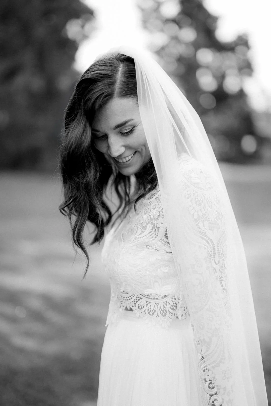 Schwarz-weiß-Foto einer glücklichen Braut in einem Spitzenkleid und einem langen Tuch, die nach unten schaut. Im Hintergrund sind unscharfe Bäume und Natur zu sehen. Die Hochzeit findet im Schloss Grünewald in Solingen bei Düsseldorf, Nähe Köln statt
