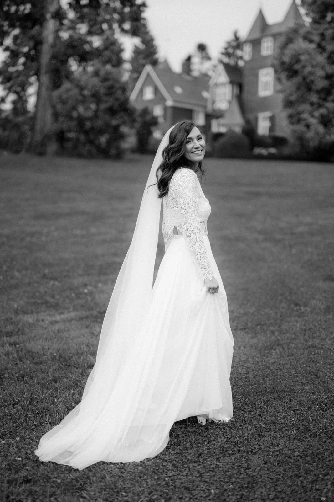 Braut in Hochzeitskleid mit Schleier im Freien auf grünem Rasen, mit Bäumen und Häuser im Hintergrund, in Schwarz-Weiß aufgenommen. Die Hochzeit findet in Solingen beim Schloss Grünewald Nähe Köln statt. 