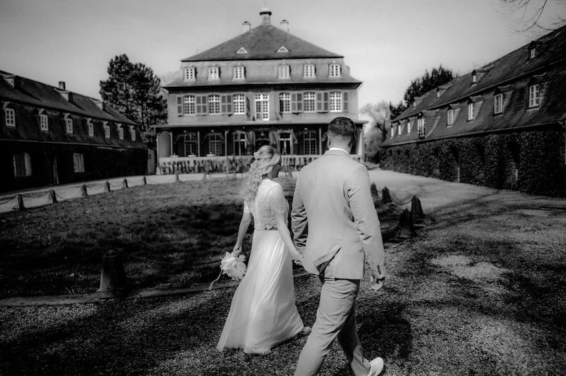 Ein Brautpaar schreitet Hand in Hand vor einem großen, mehrstöckigen Gebäude bei Sonnenlicht. Die Hochzeit findet im Schloss Eicherhof in Leichlingen bei Düsseldorf, Nähe Köln statt. 