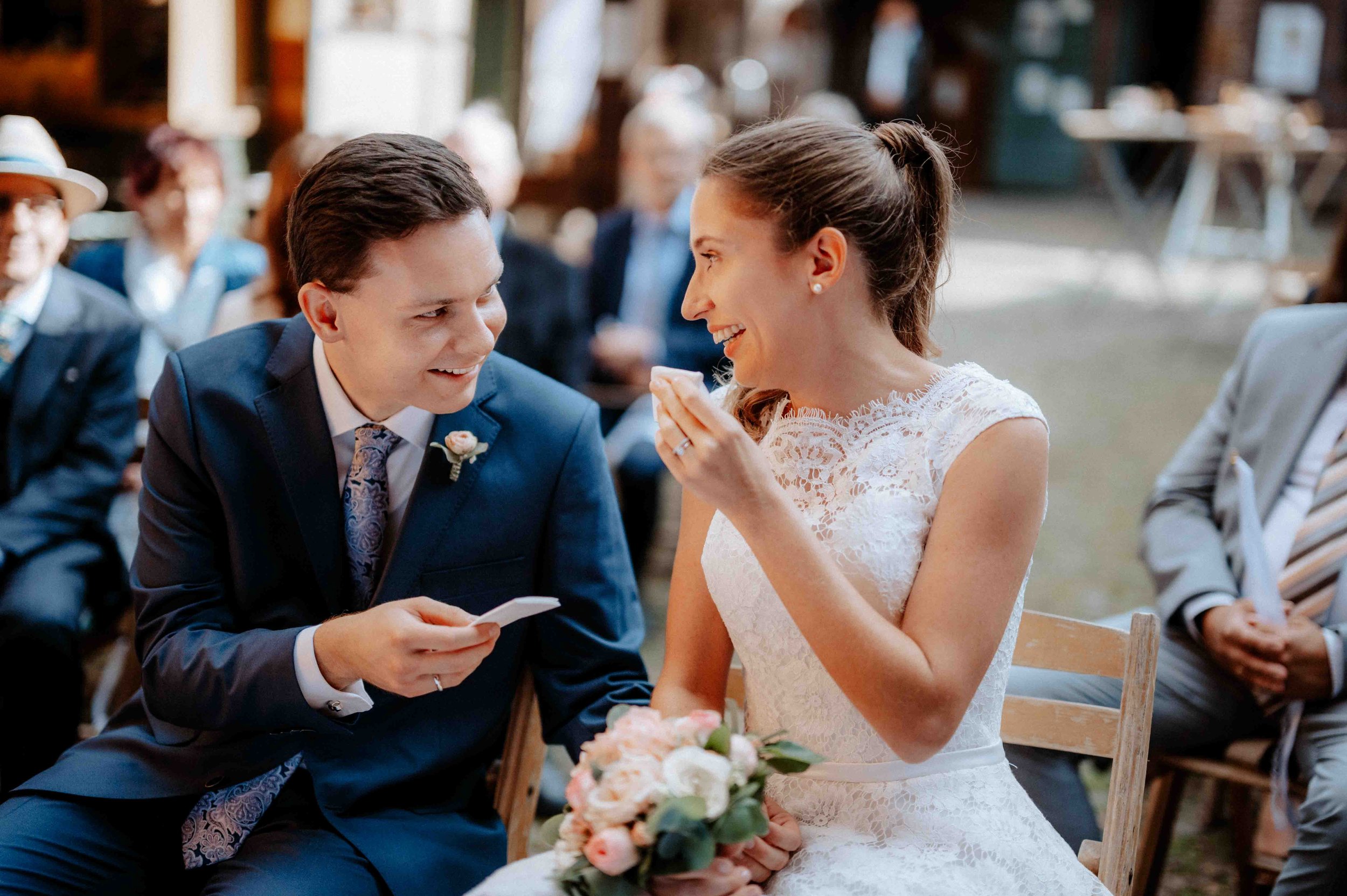 Ein Brautpaar bei ihrer Hochzeit, der Bräutigam lächelt, während die Braut lachend eine Hand vor den Mund hält, im Hintergrund sitzen Gäste bei einer Zeremonie. Die Hochzeit findet in Düsseldorf, Nähe Köln statt. 