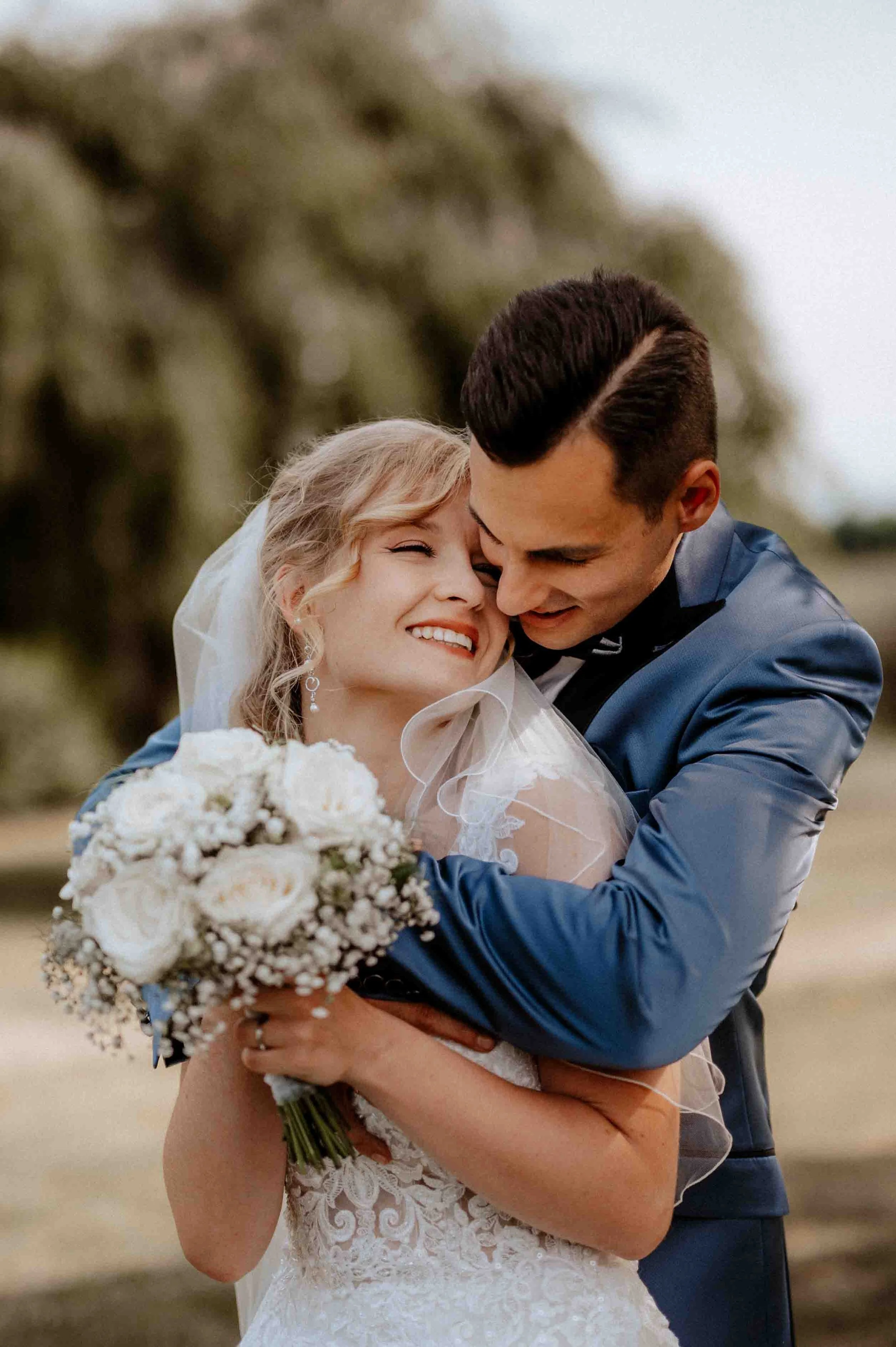 Ein Brautpaar umarmt sich liebevoll im Freien, sie hält einen Brautstrauß aus weißen Blumen, sie trägt ein weißes Hochzeitskleid mit Spitzen, er trägt einen dunklen Anzug. Die Hochzeit findet in Düsseldorf, Nähe Köln statt. 