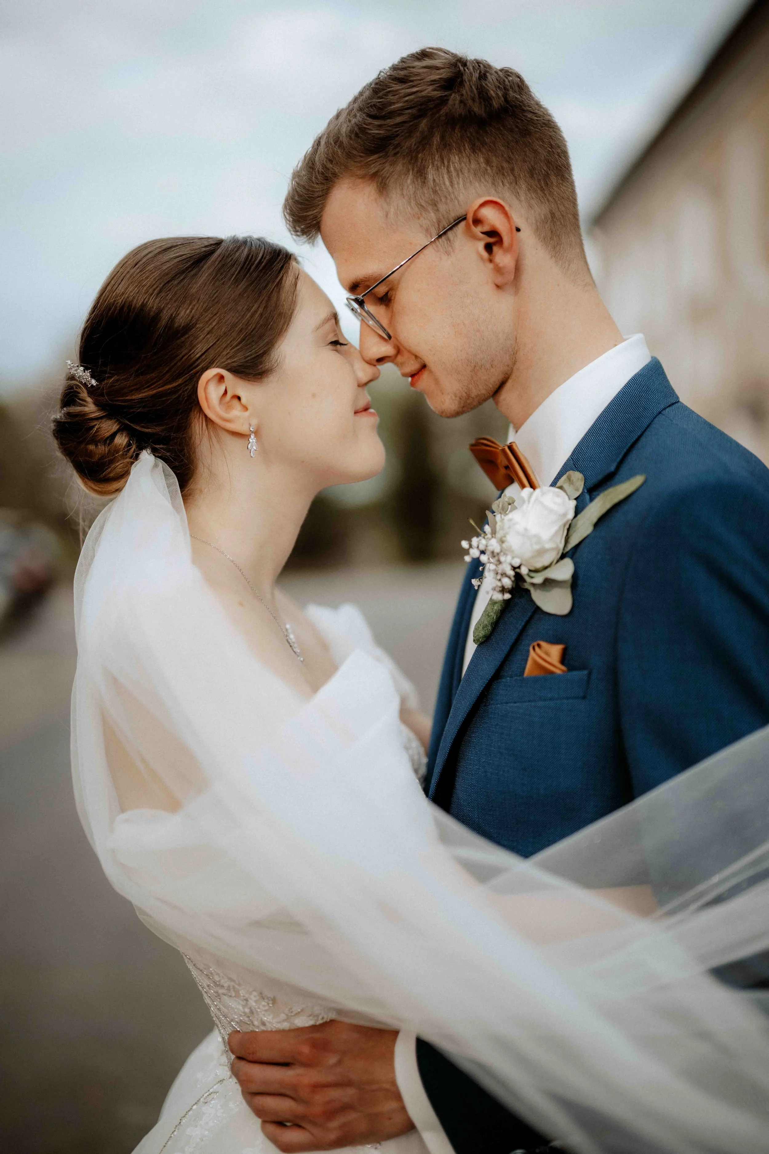 Ein Brautpaar steht sich bei einer Hochzeit sehr nahe mit den Stirn aneinander, lächelt und berührt sich. Sie trägt ein Brautkleid mit Schleier, er einen Anzug mit Fliege und Rosenboutonnière. Die Hochzeit findet in Düsseldorf, Nähe Köln statt. 