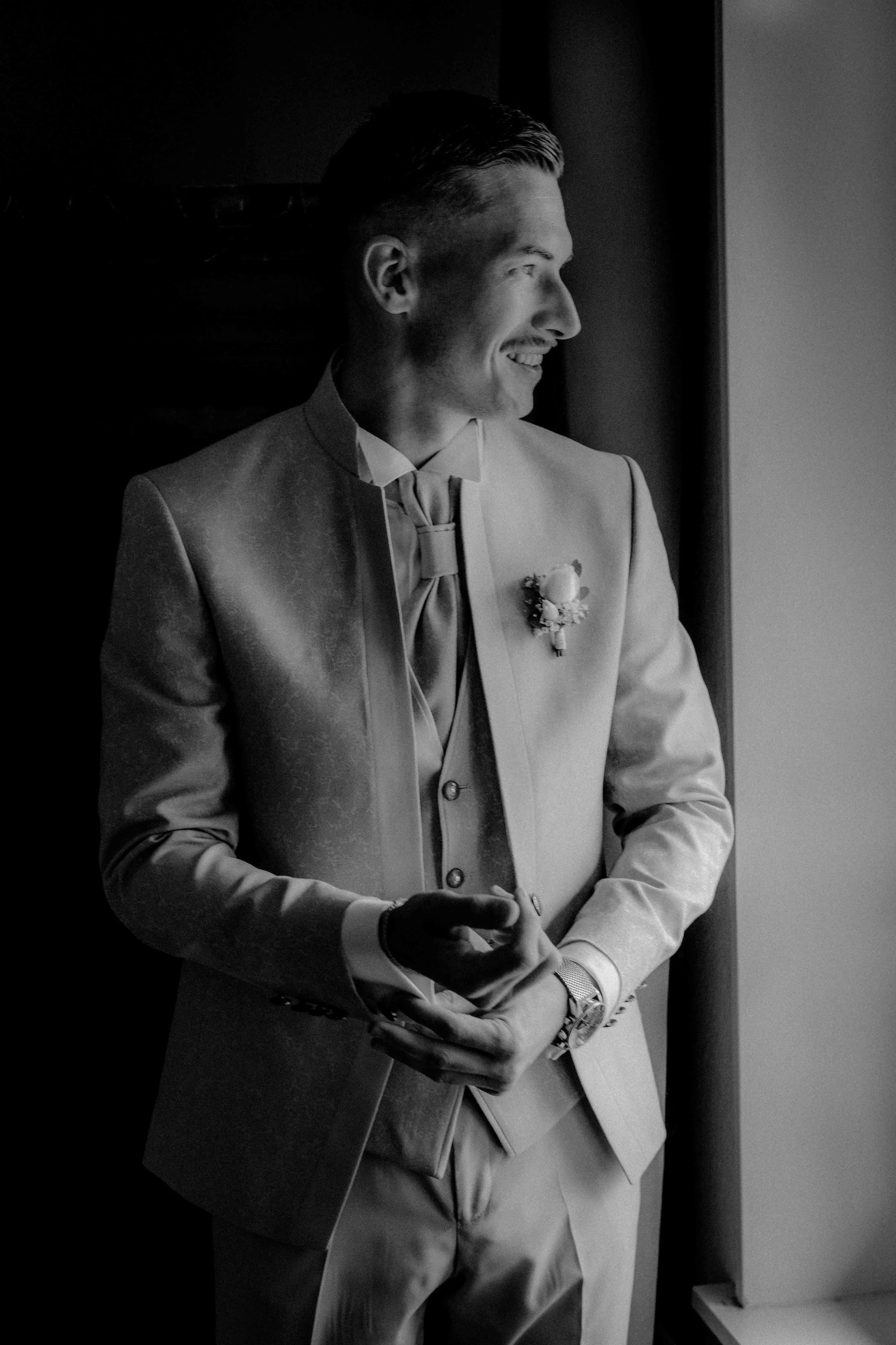 Mann in Hochzeitsanzug steht am Fenster, lächelt und schaut nach draußen. Die Hochzeit findet im Landgut Stüttem in Wipperfürth, Nähe Köln statt. 