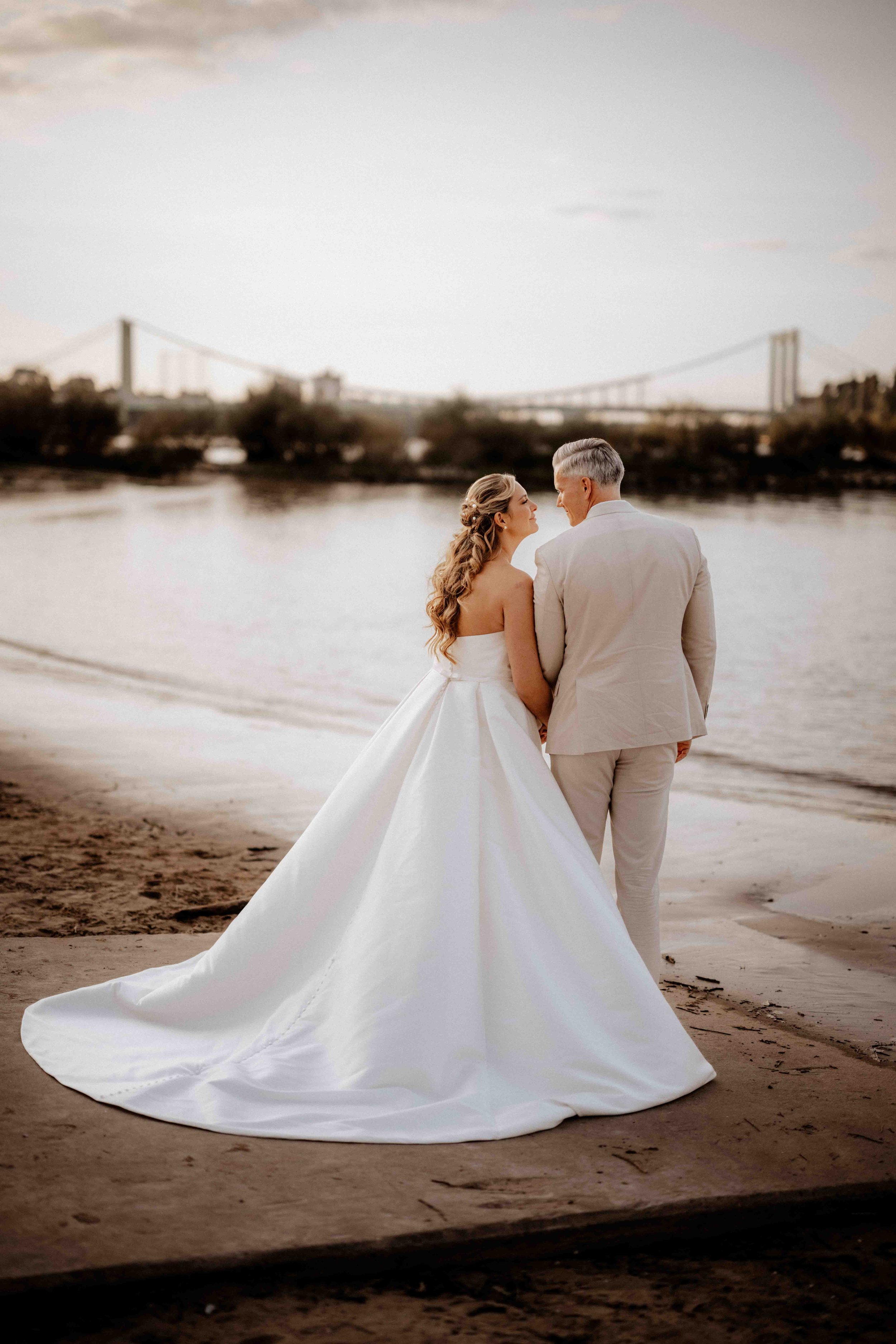 Ein Brautpaar steht am Flussufer mit Blick auf eine Brücke im Hintergrund, umgeben von Natur, bei Sonnenuntergang. Die Hochzeit findet in Köln Rodenkirchen am Rhein statt. 