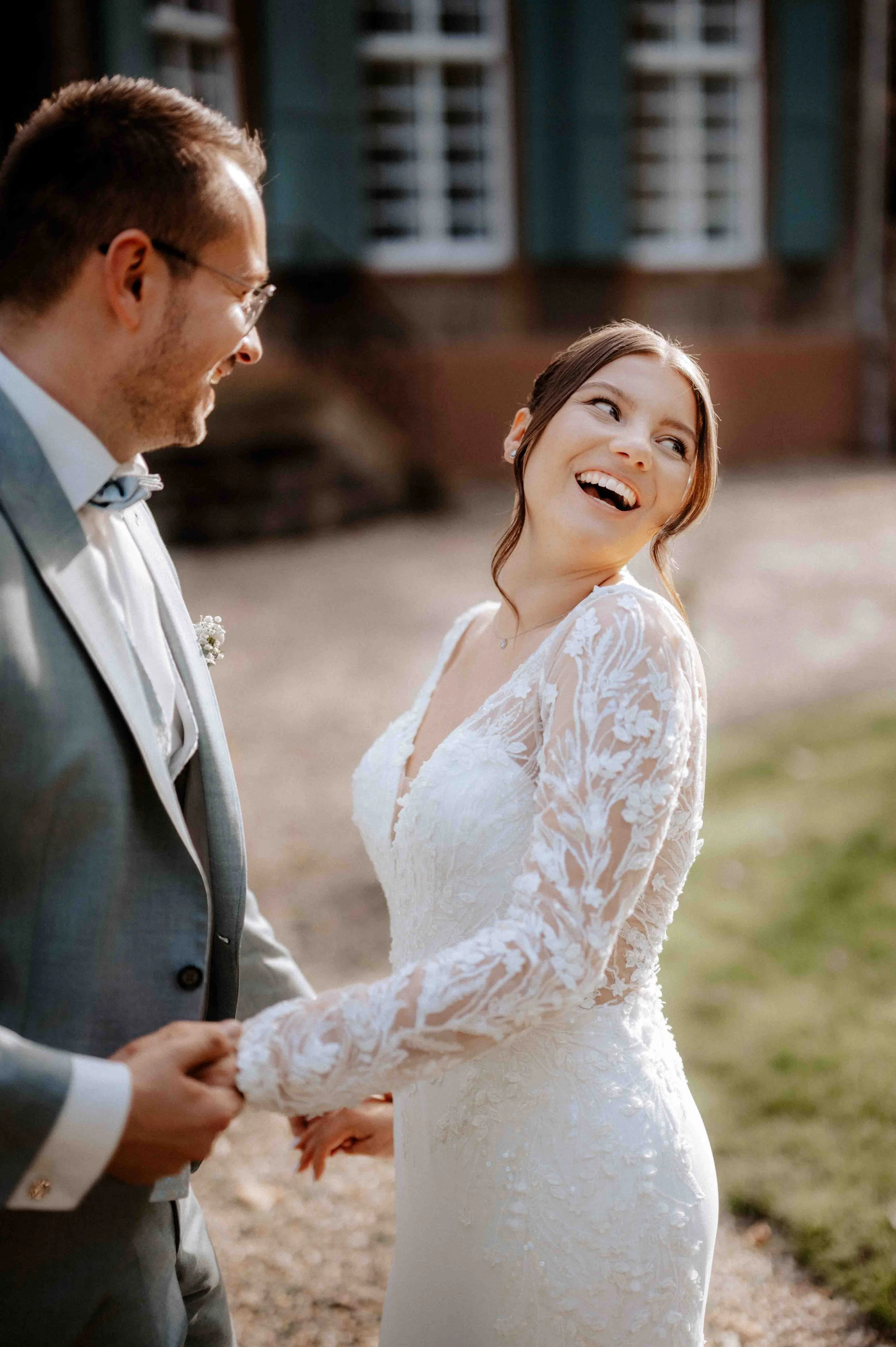 Ein glückliches Brautpaar hält Händchen bei der Hochzeit im Freien, die Braut trägt ein weißes Spitzenkleid, beide lachen. Hinter ihnen ist ein Gebäude mit blauen Fensterläden. Die Hochzeit findet im Schloss Eicherhof in Leichlingen bei Düsseldorf, N