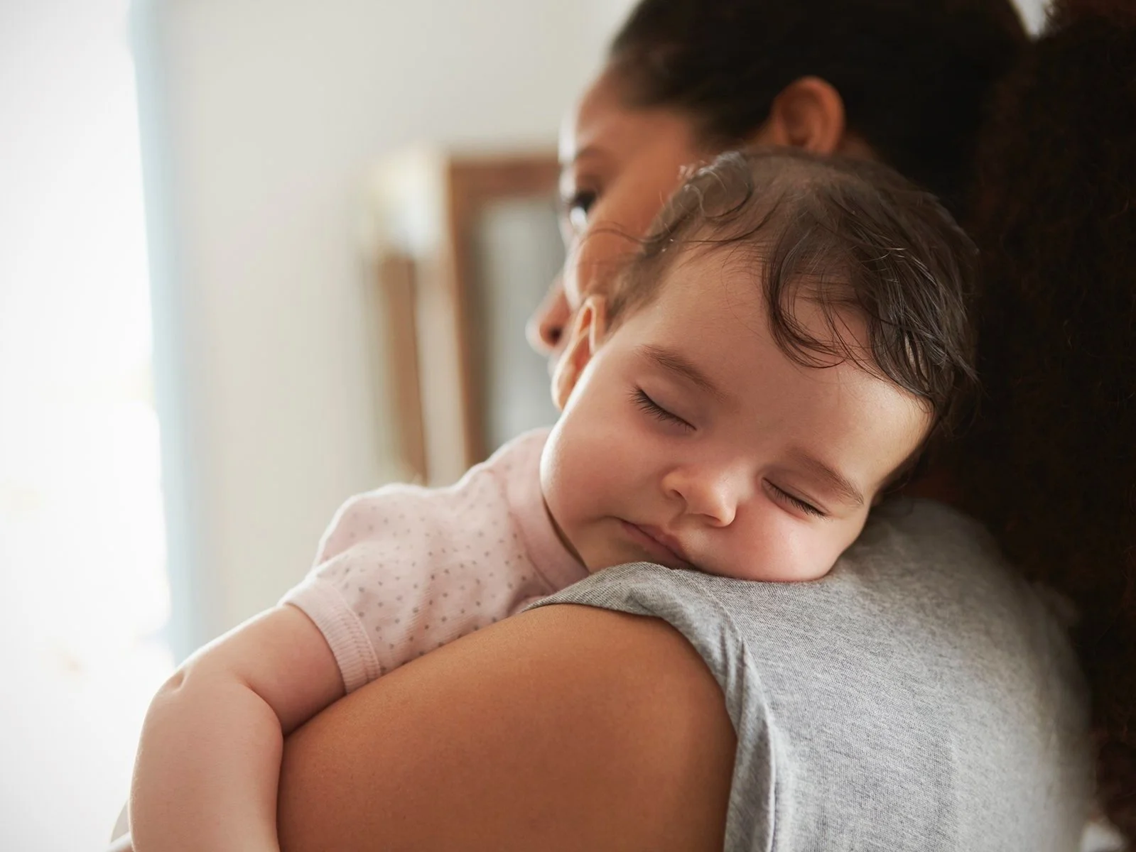 Close-up portrait of baby sleeping with their head resting on their mother’s shoulder.