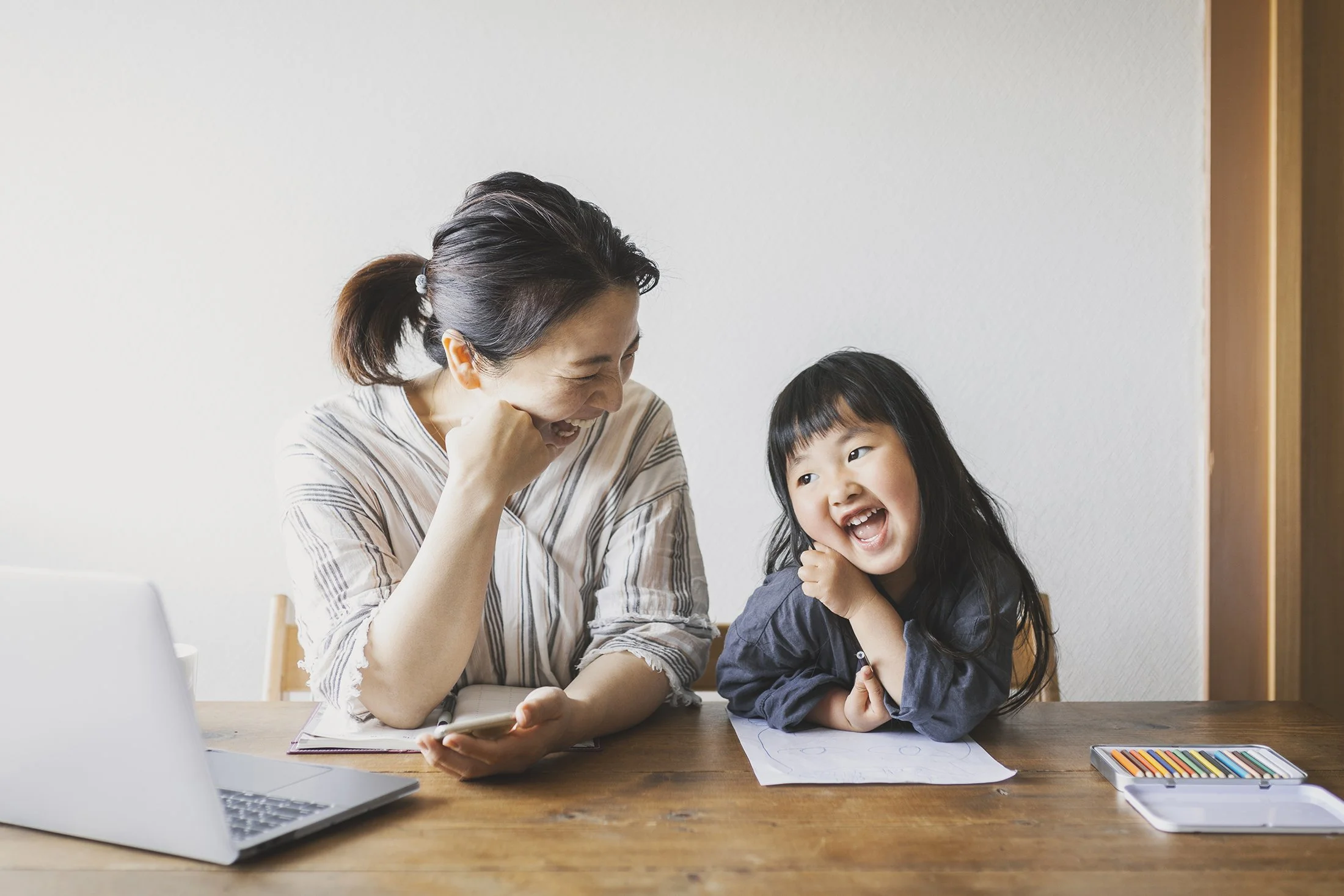 Mother and daughter sitting together at a table, smiling at each other. The mother is working on a laptop computer, the child is drawing with colored pencils.