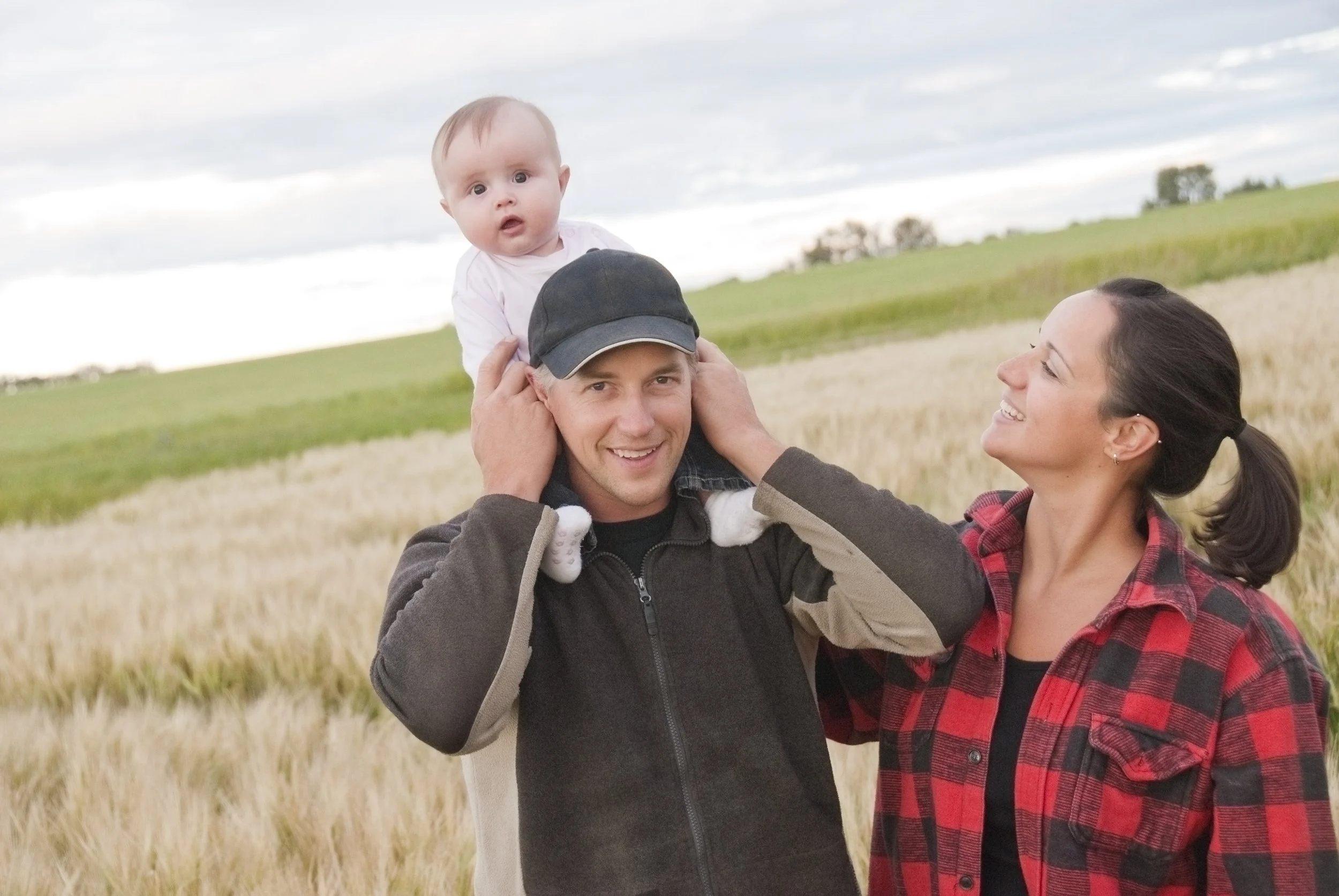Family standing together in a rural setting. The father is holding their baby on his shoulders.