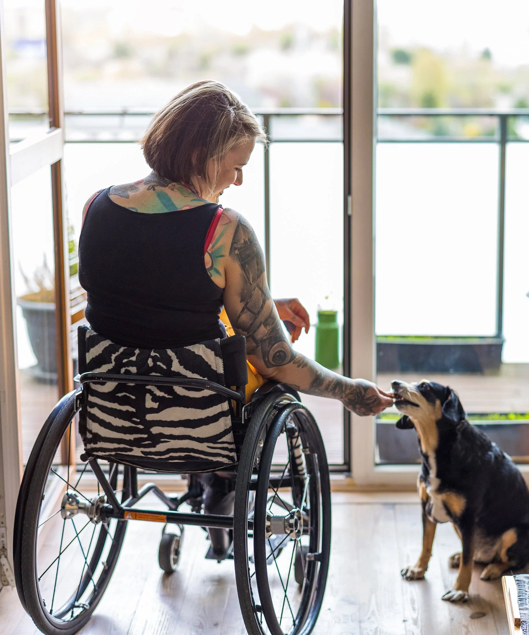 Young woman with tattoos on her arms sitting in a wheelchair looking out the window. She is smiling and feeding a treat to her dog.