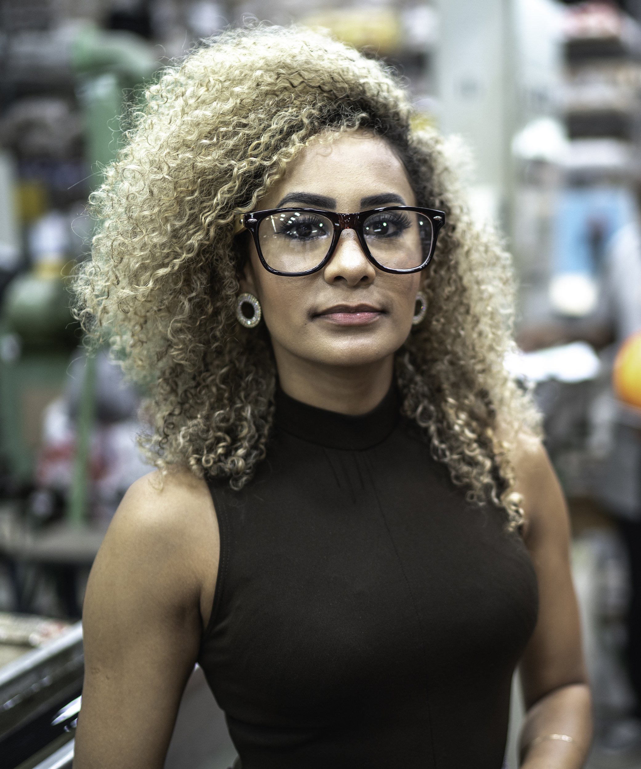 Portrait of a young, smiling woman with bleached-blond coiled hair and stylish thick-rimmed glasses.