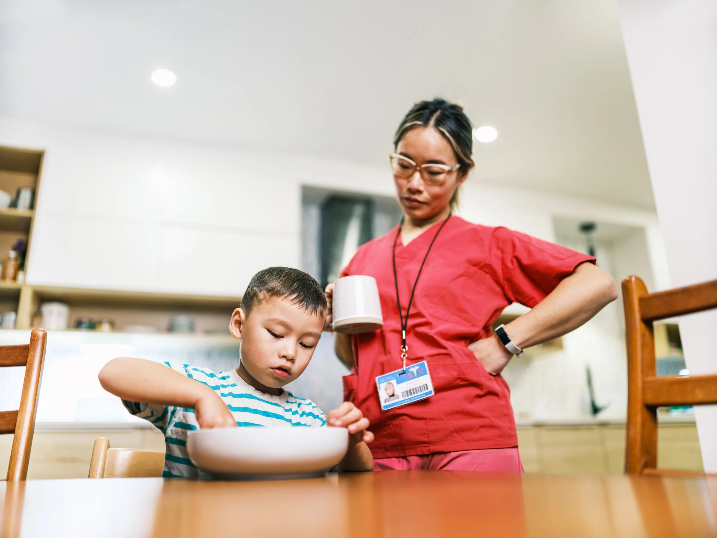 A young boy is sitting at a table and mixing something in a bowl. A woman in a red uniform is standing behind him, holding a mug.