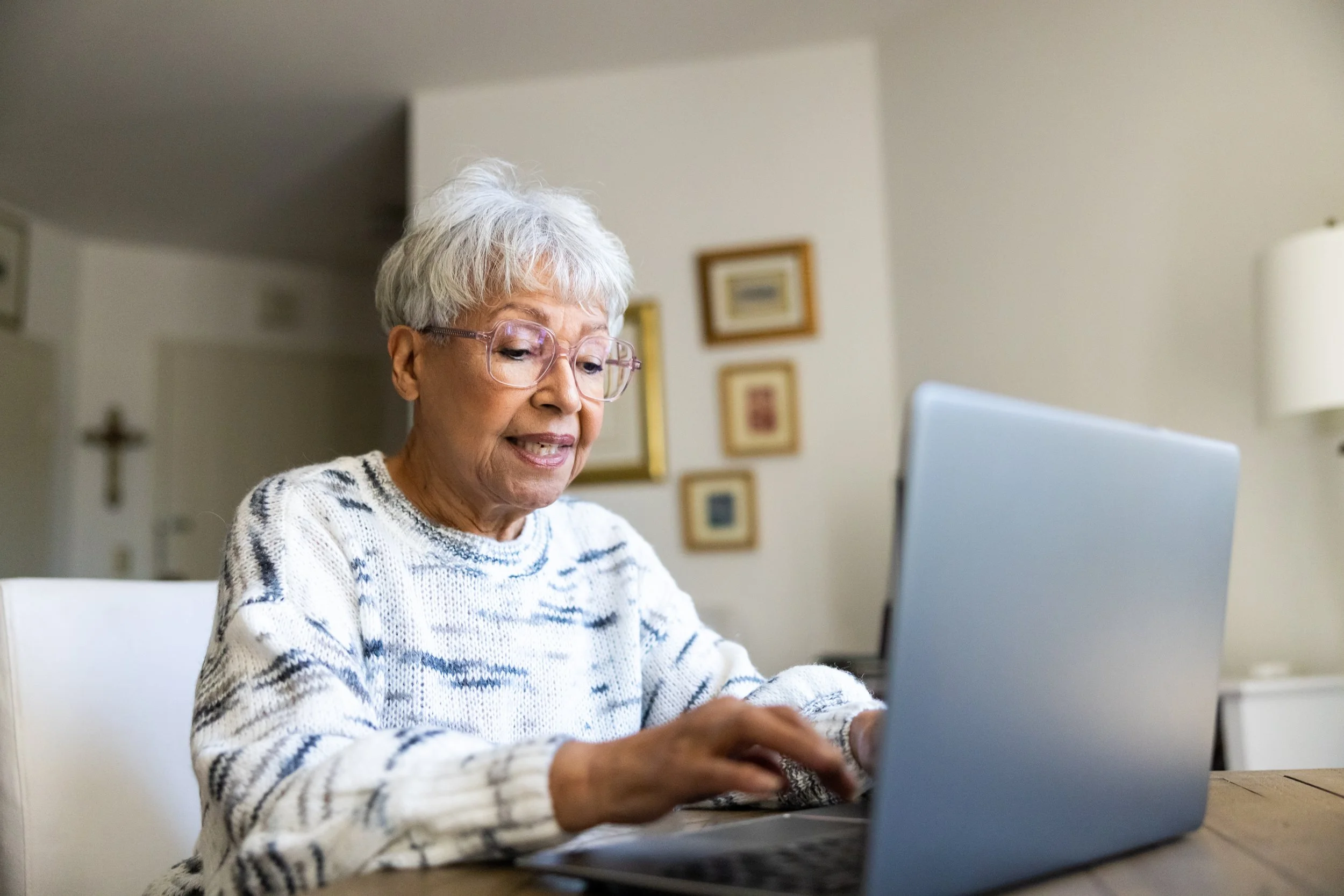 An elderly woman with glasses is using a laptop. She is sitting at a table typing on a laptop.