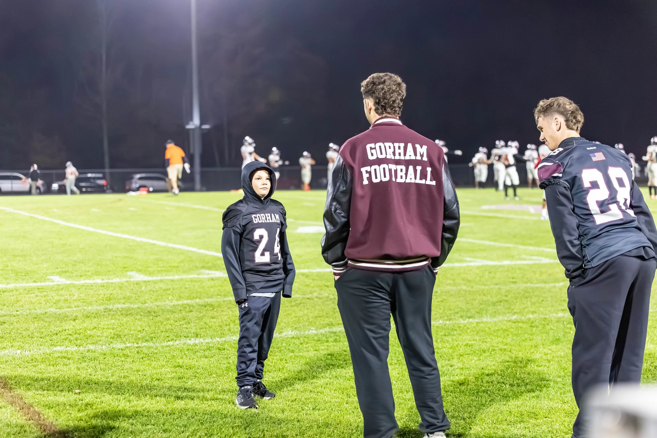 Football players and a young fan on a football field at night, with players wearing maroon and navy jerseys, and the fan wearing a black hoodie with 'Gorham' and '24' on it, engaging in conversation.