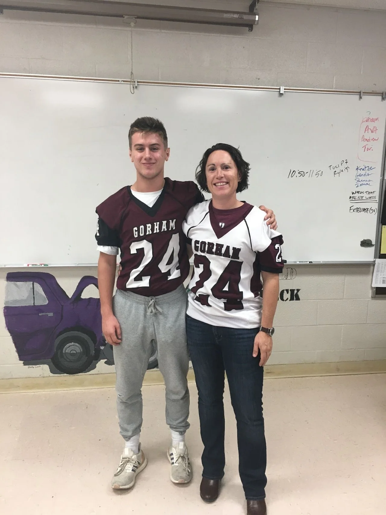 A teenage boy and a middle-aged woman standing together in a classroom, both wearing maroon and white football jerseys that say 'Gorham' and the numbers 24 and 24, smiling at the camera.