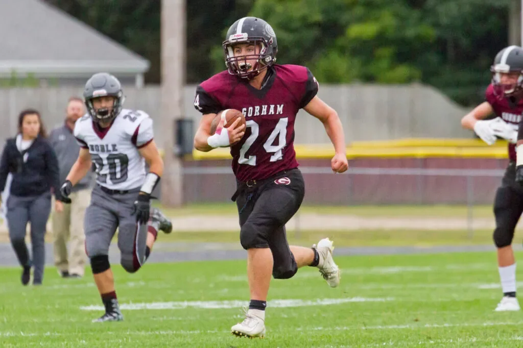 A football player from Gorham runs with the ball while a Boone player chases behind him during a game on a grassy field.