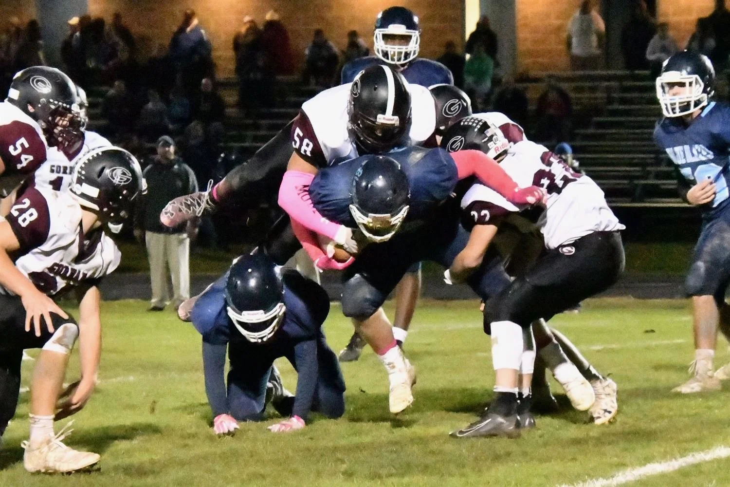 American football game with players tackling and blocking on a grassy field at night, spectators in the background.