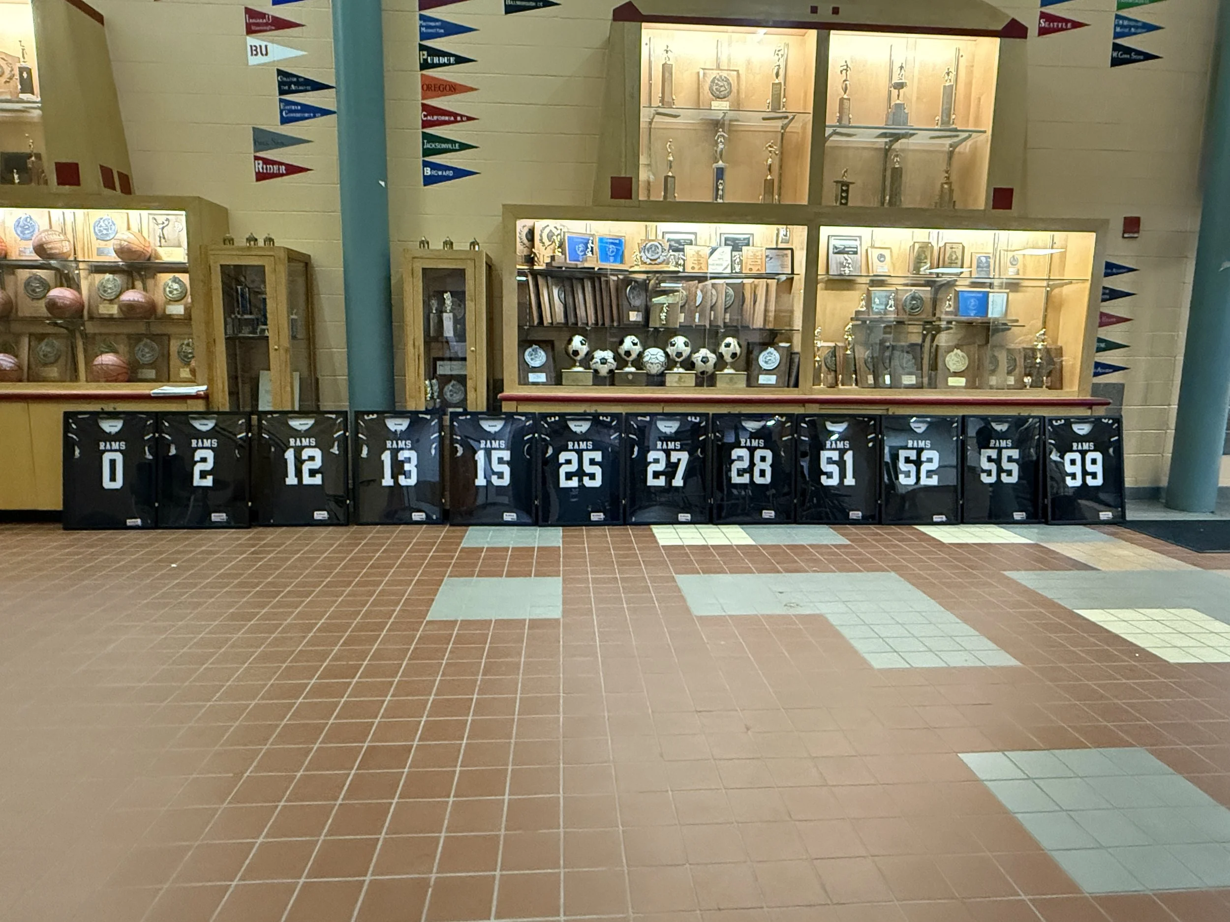 Display of framed sports jerseys, trophies, medals, and soccer balls inside a glass case at a school or sports hall, with flag banners hanging on the wall.