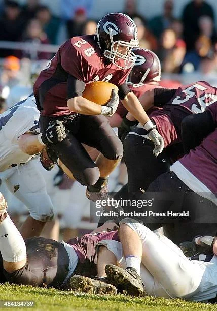 A football player in maroon and black uniform jumping over players on the ground during a game.