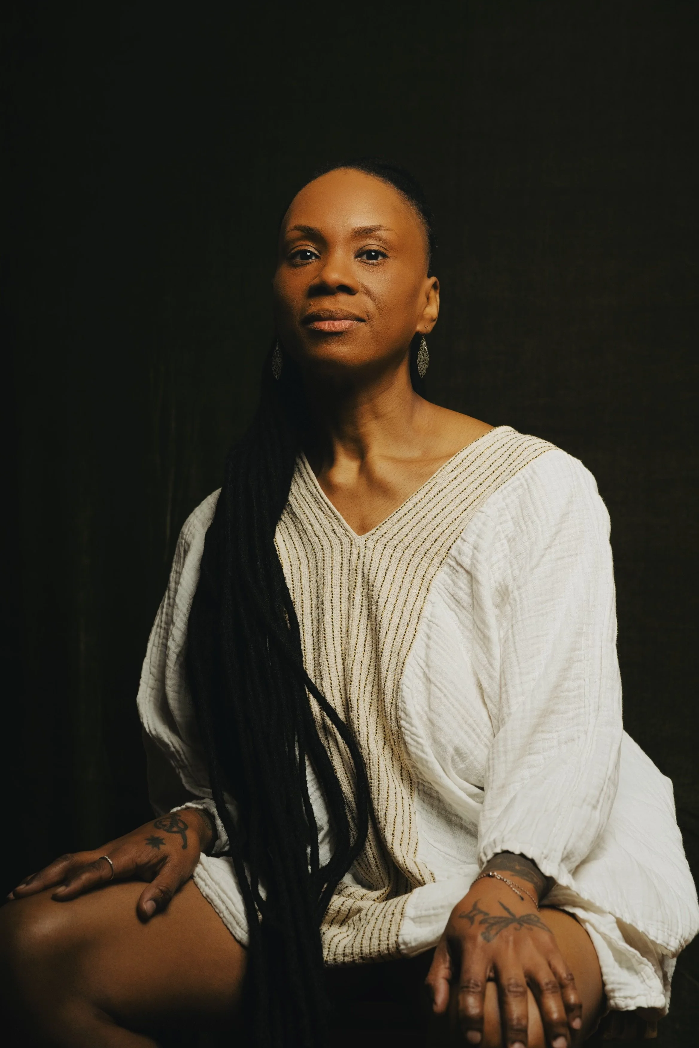 A woman with long black hair, earrings, and tattoos on her hands, sitting against a dark background, wearing a white textured blouse.