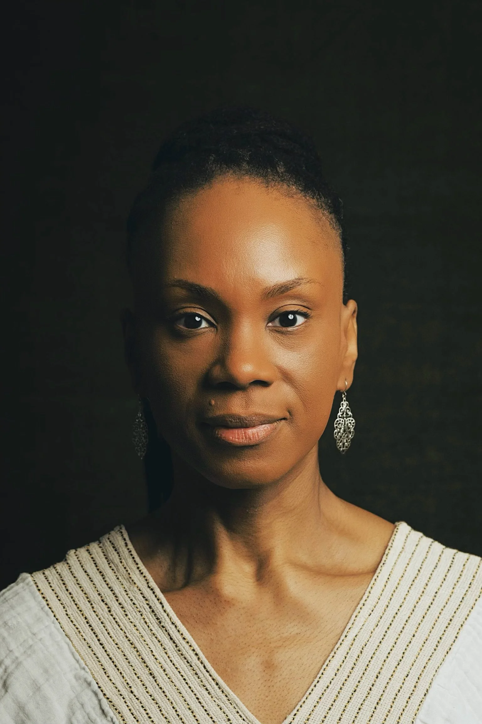 Close-up portrait of a woman with dark skin and short, textured hair, wearing silver earrings and a white blouse with gold embroidery, looking directly at the camera against a dark background.