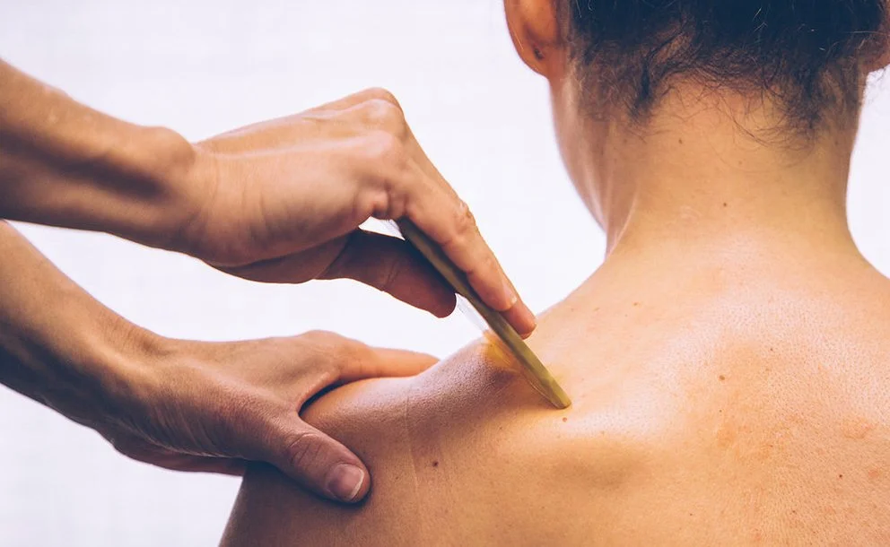 Person using a wooden tool to perform gua sha therapy on a woman's shoulder.