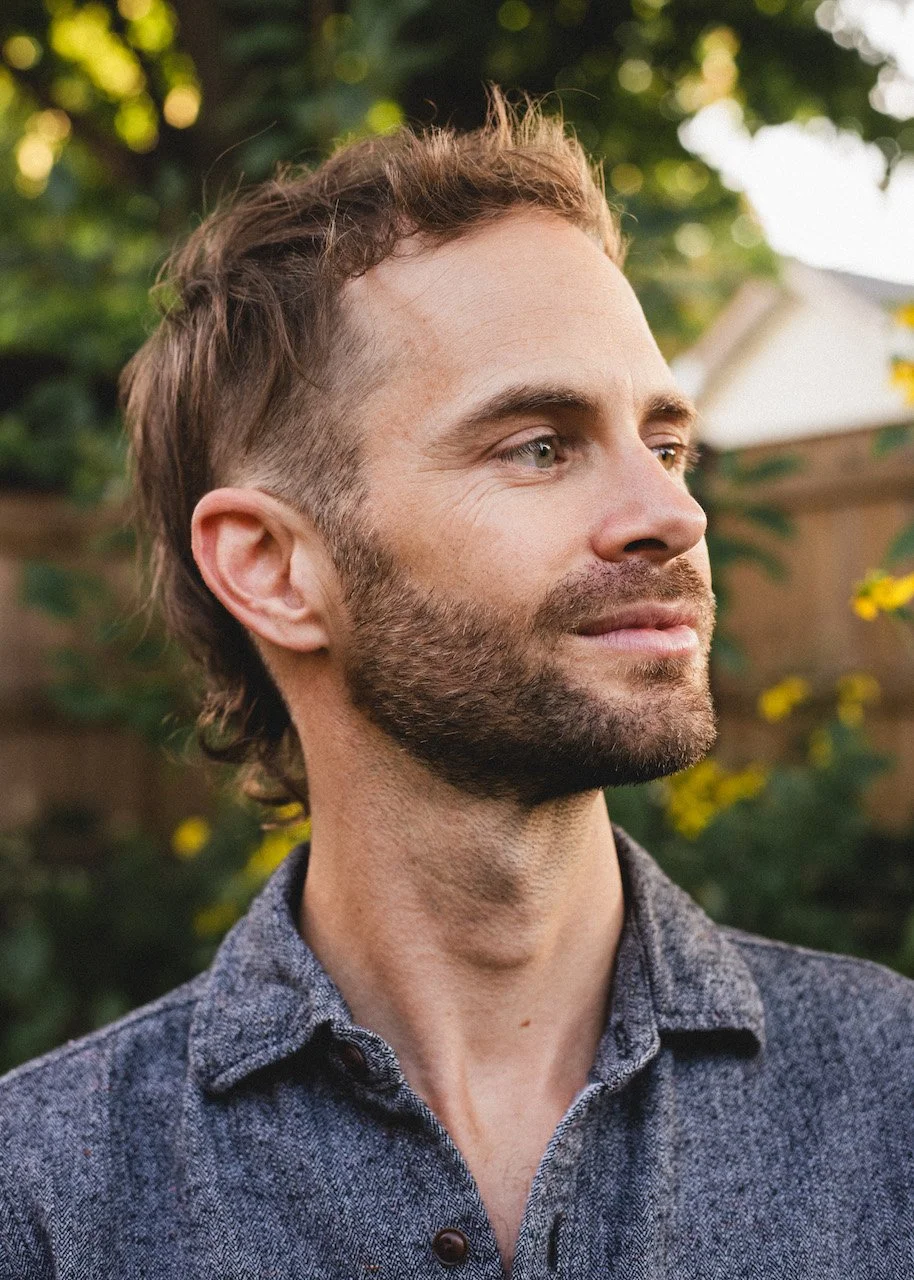 Side profile of a man with a beard and short brown hair, wearing a gray button-up shirt, outdoors with green foliage and a house in the background.