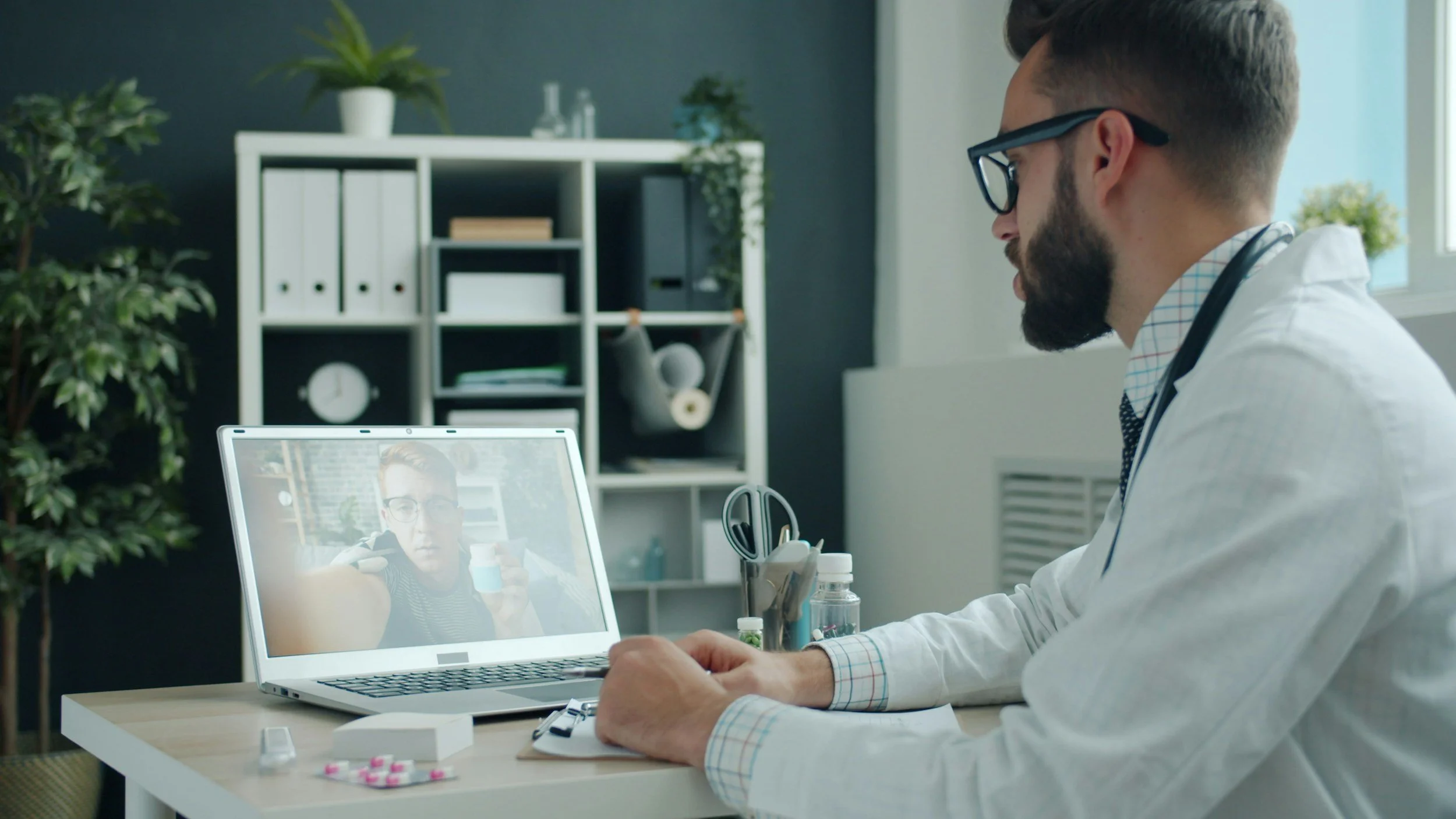 Doctor sitting at desk, taking notes, talking to a patient on a video call on laptop, in a well-lit office with plants and shelves.