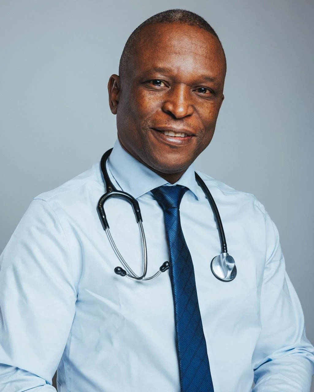 Portrait of a smiling male doctor with a stethoscope around his neck, wearing a white shirt and a blue tie, against a neutral background.