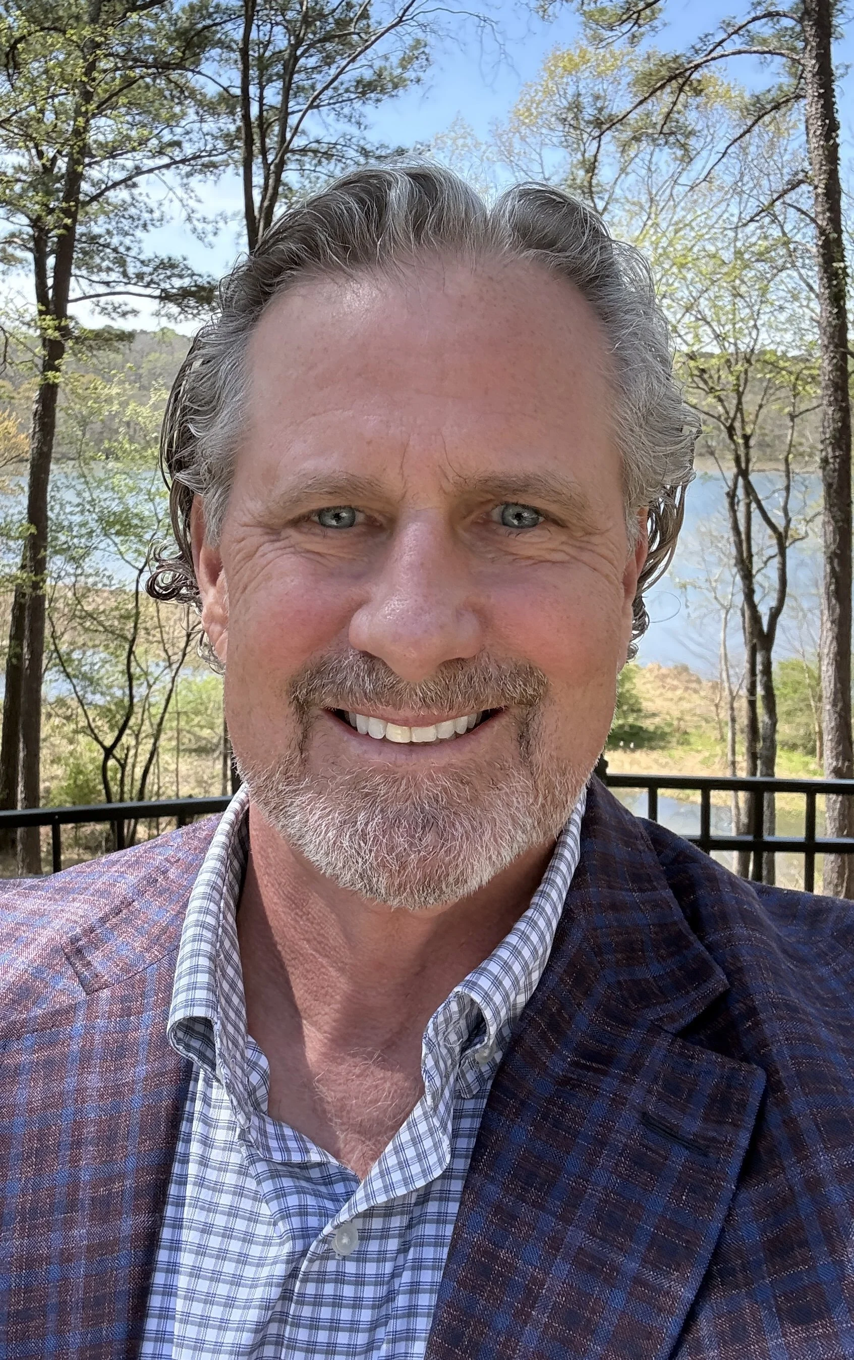 A man with gray hair and beard smiling outdoors, with a lake and trees in the background.