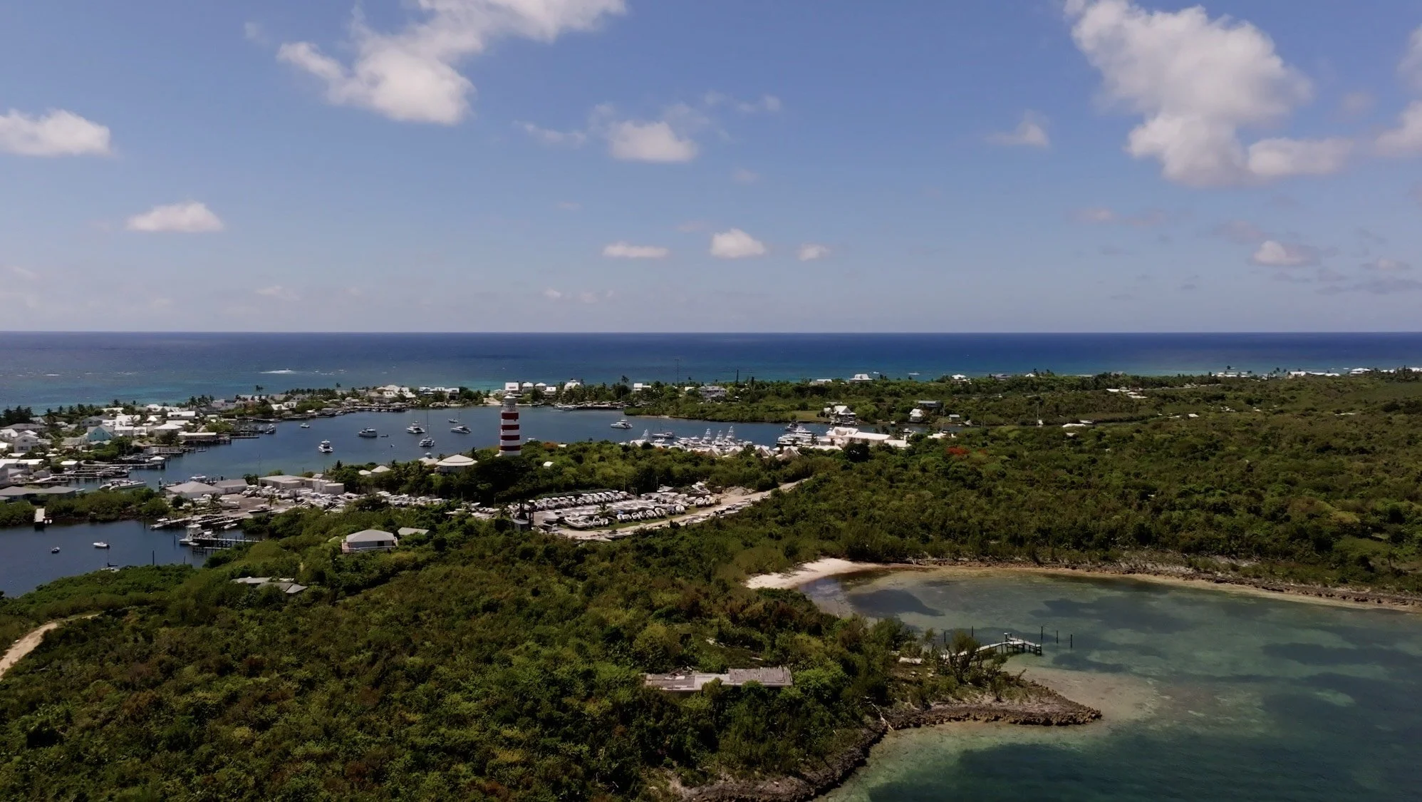 Aerial view of a coastal area with a marina, small boats, lush greenery, a lighthouse, and the ocean in the background under a partly cloudy sky.