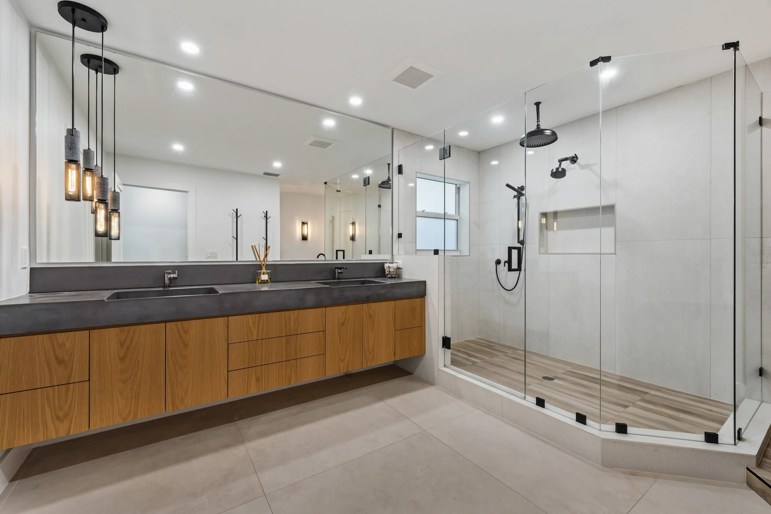 Modern bathroom with a double vanity with a dark gray countertop, wooden cabinets, a large mirror, and pendant lights. There is a glass-enclosed walk-in shower with black fixtures, a rainfall showerhead, a window, and a built-in shelf. The bathroom h