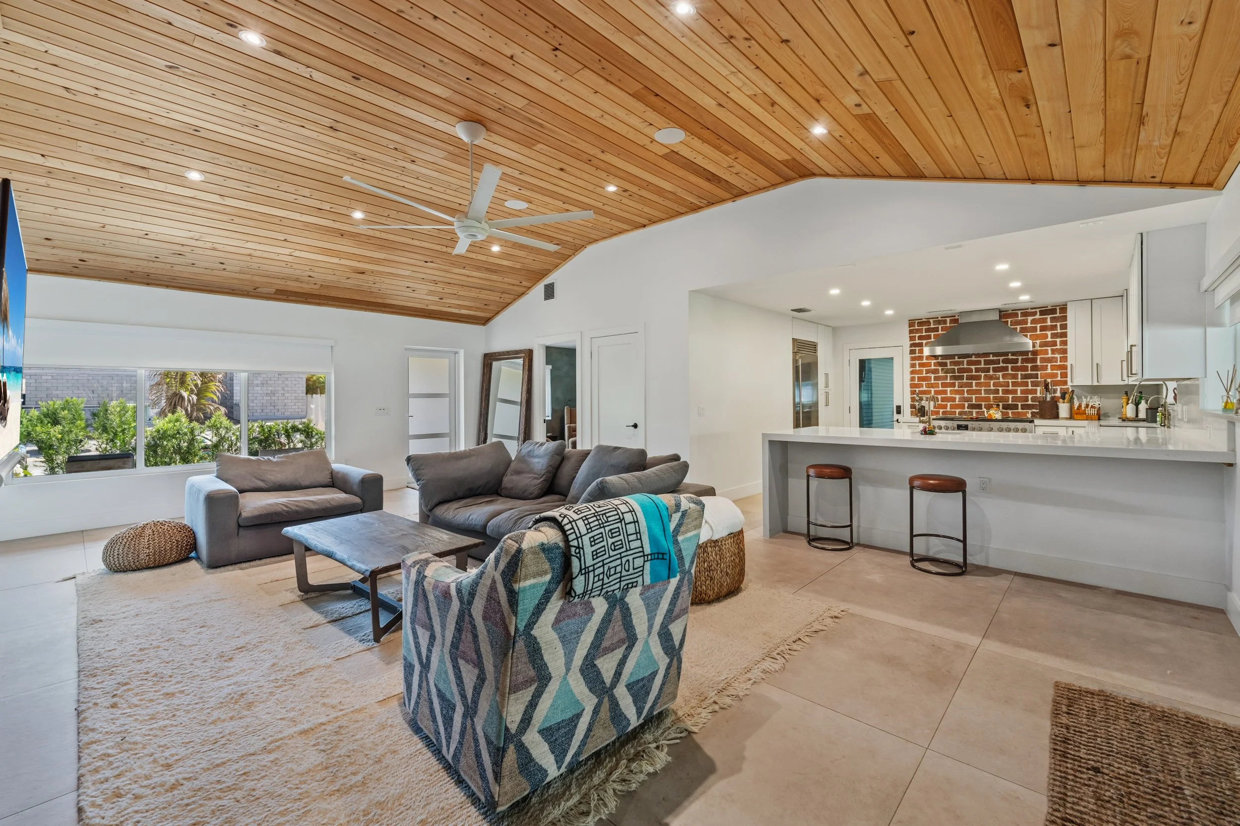 Open concept living room and kitchen with a vaulted wooden ceiling, large window, and neutral furniture, including a sofa, armchair, and coffee table, with a brick accent wall in the kitchen area.