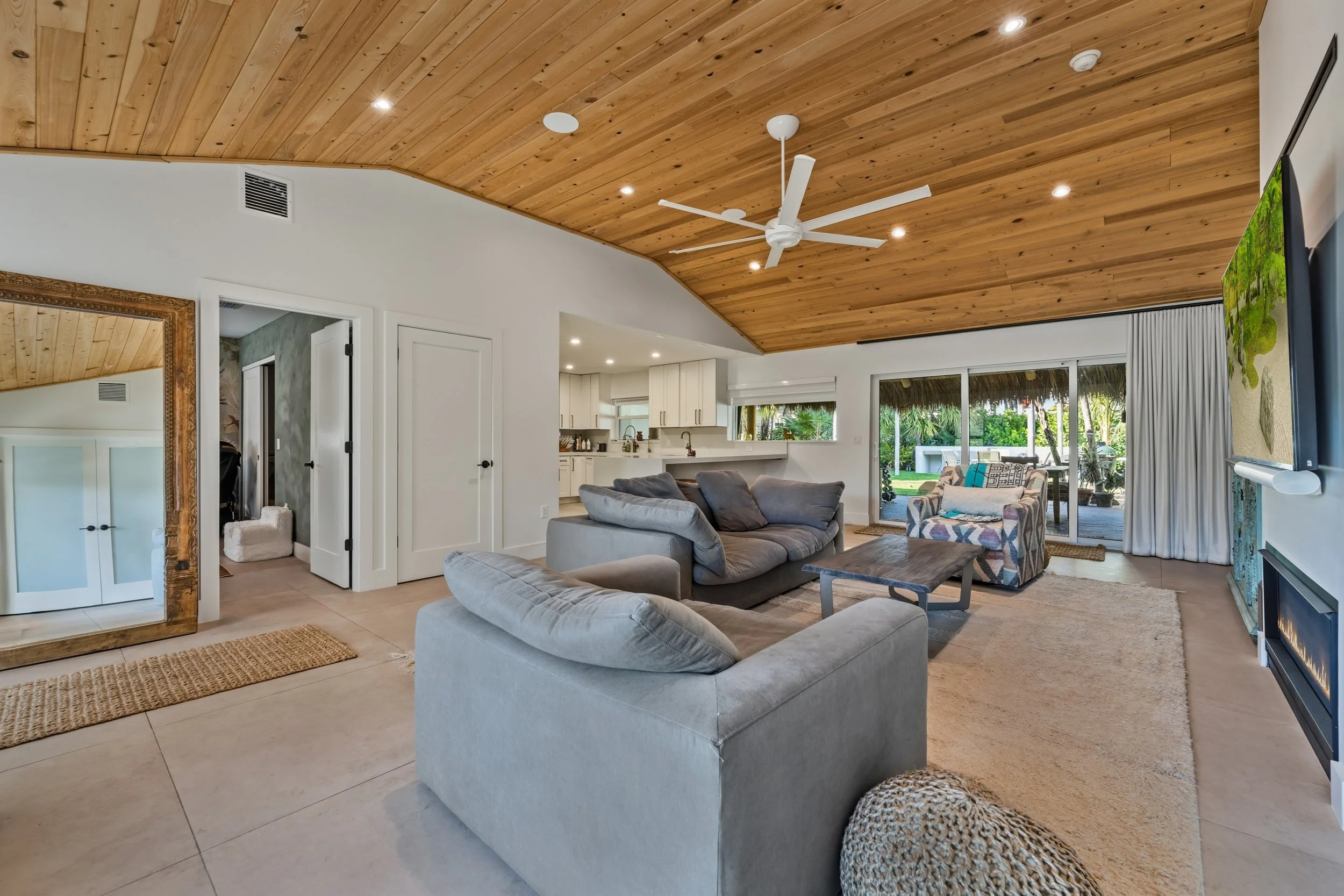 Living room with a vaulted wood ceiling, white walls, gray and patterned sofas, a coffee table, a large mirror, a fireplace, and sliding glass doors leading to an outdoor patio with trees and grass.