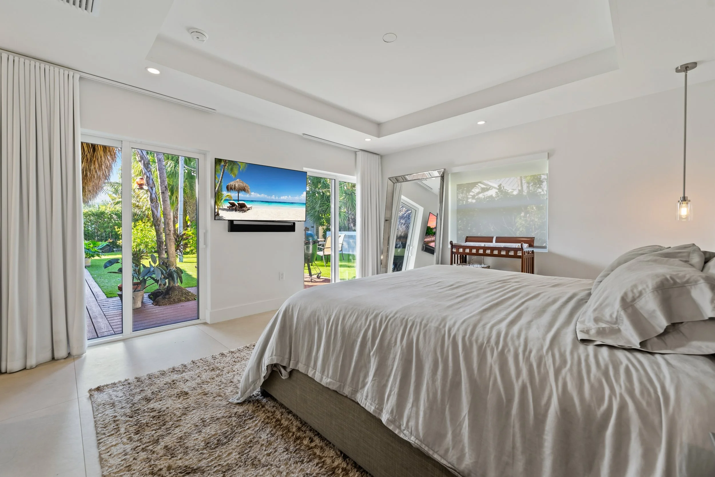 Modern bedroom with large bed, sliding glass door to outdoor patio, TV on wall, mirror, and tropical garden view.